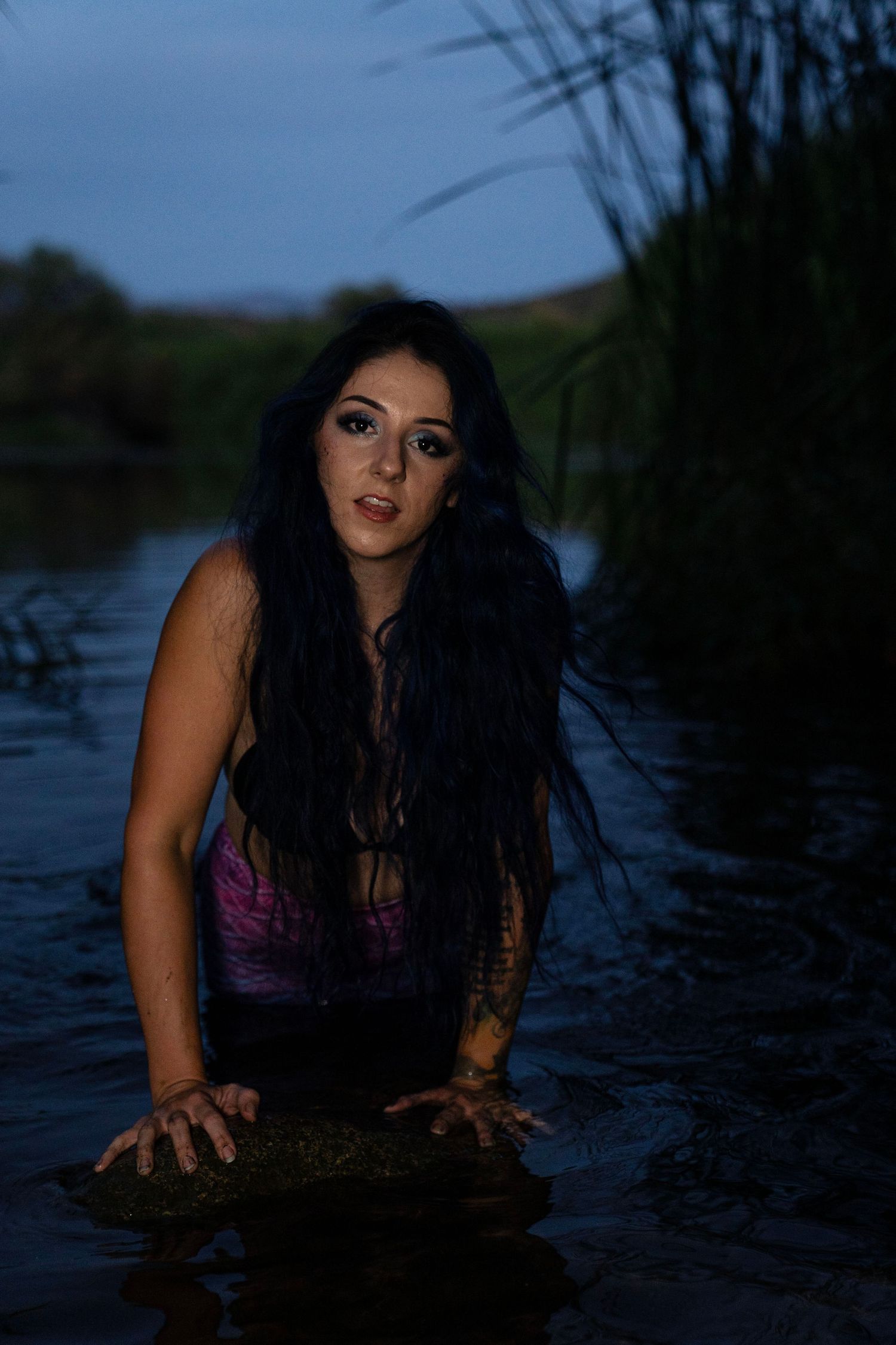 Dark-haired woman in purple top emerging from a river at dusk, surrounded by tall reeds.