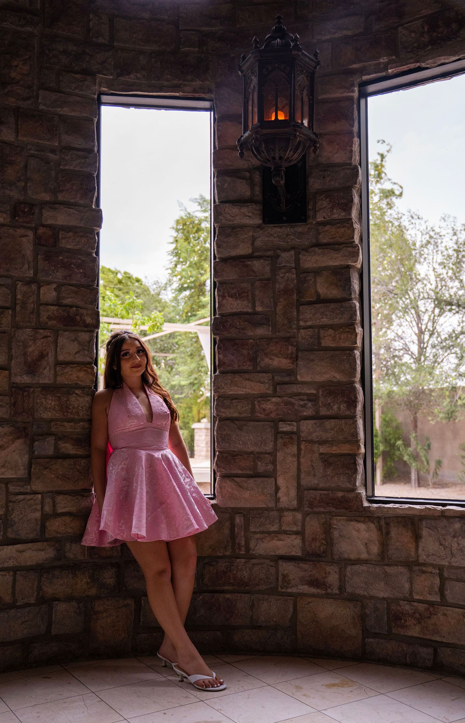Young woman in pink dress posing by stone wall windows with vintage lantern.