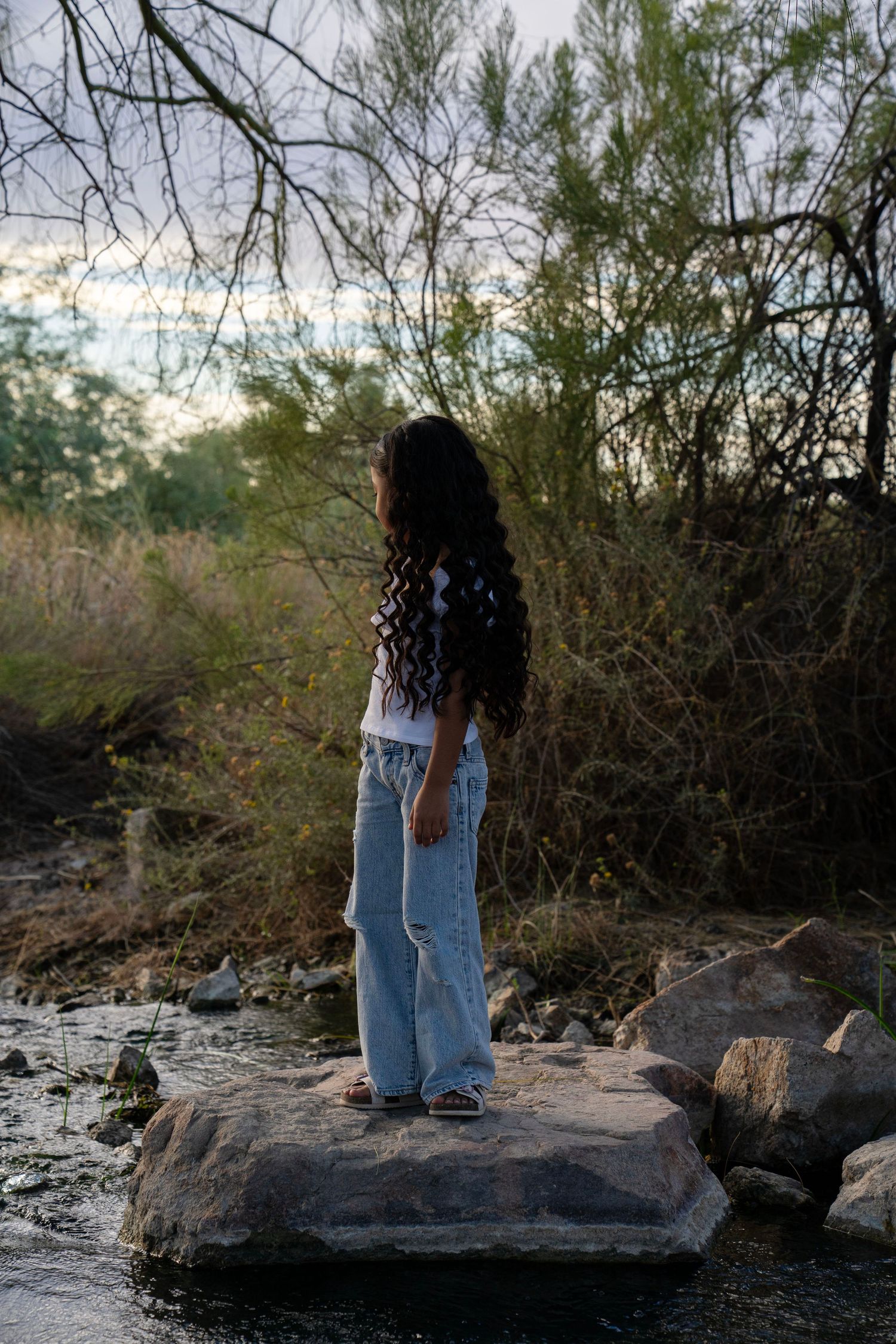 A girl with long curly hair stands on a rock by a stream, wearing wide-leg jeans, surrounded by trees.