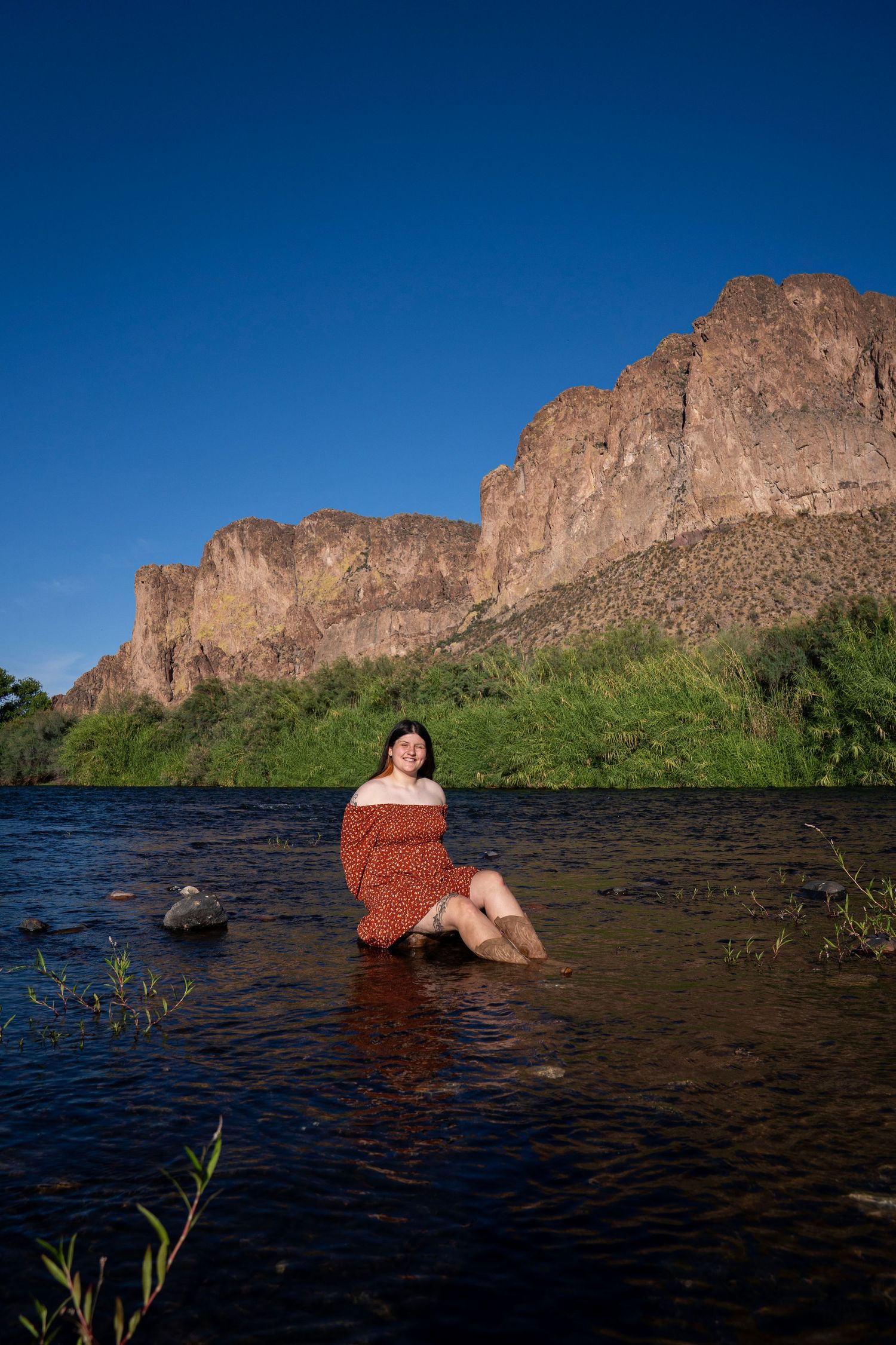 Woman in red floral dress sitting in a river with dramatic rocky cliffs and blue sky in the background.