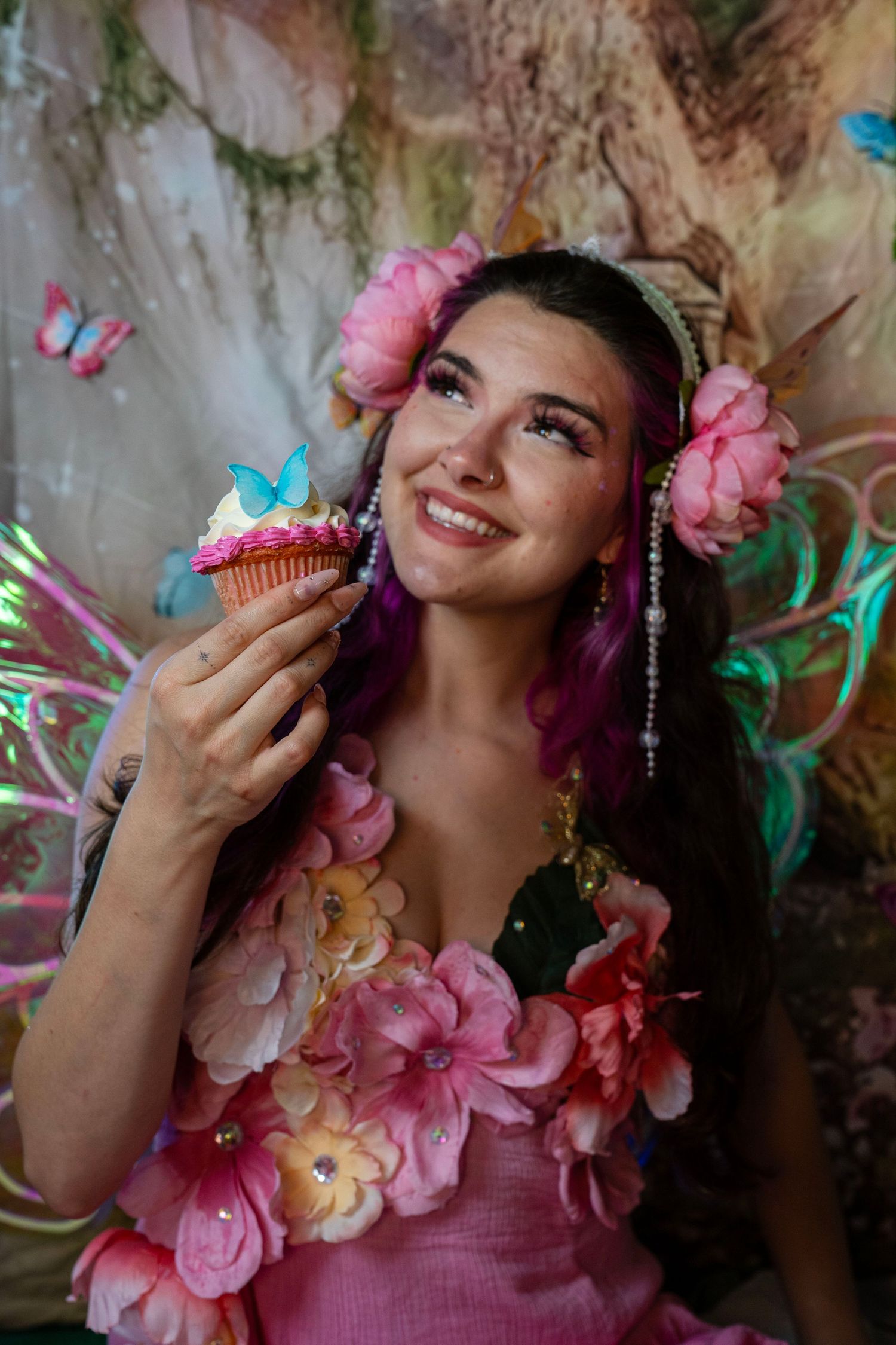 Smiling woman dressed as a fairy with pink flowers and butterfly wings holds a cupcake in a magical forest. Fantasy Photoshoot