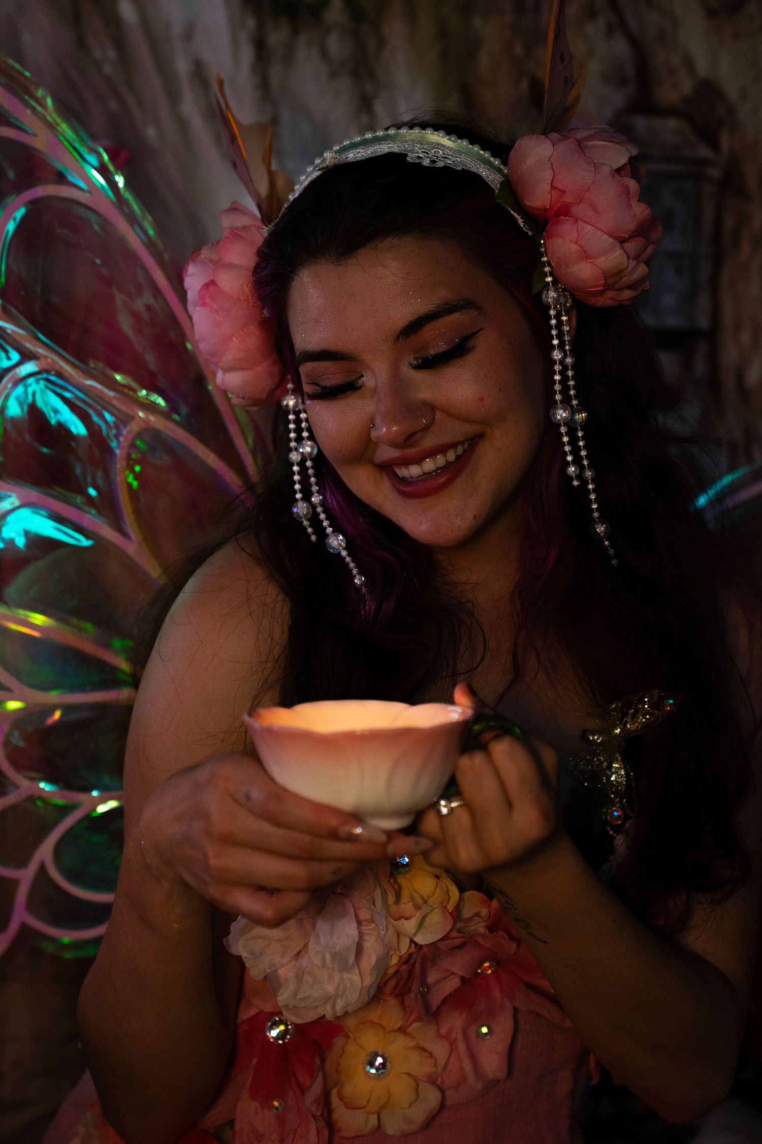 Smiling woman in floral fairy dress holds glowing lotus candle with colorful iridescent wings behind her. Fantasy Photoshoot