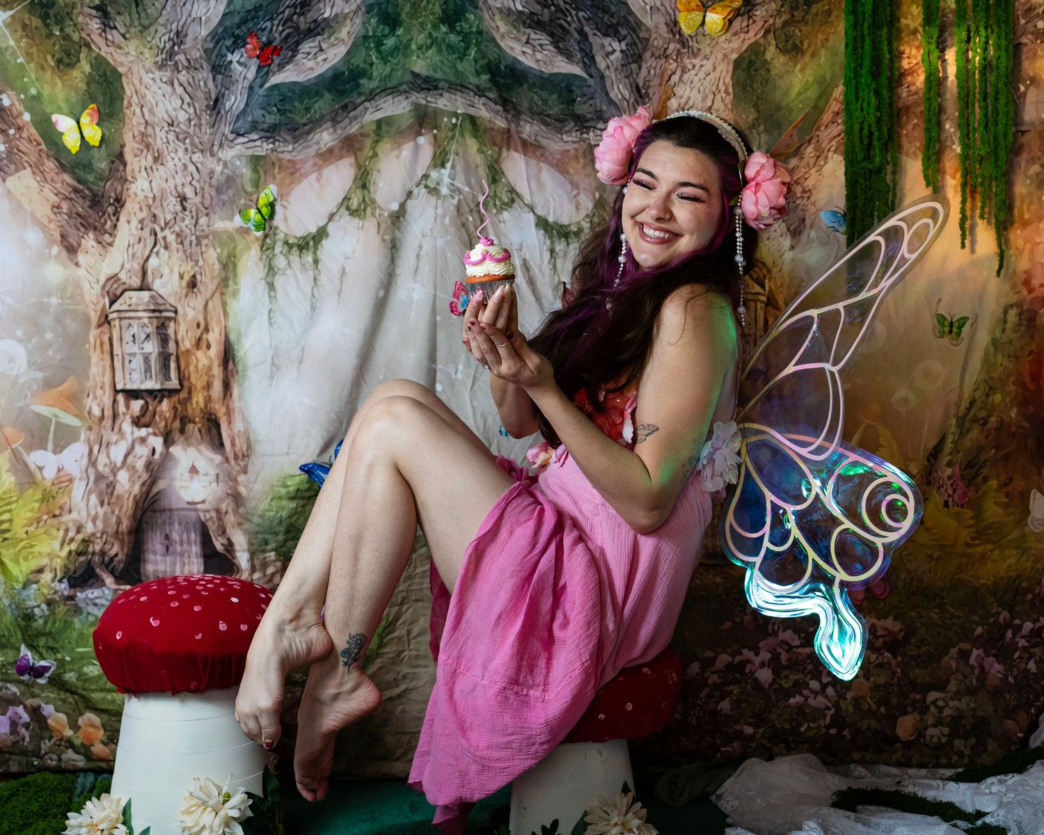 Smiling woman in pink fairy dress with holographic wings sits on mushroom holding cupcake in fantasy woodland scene. Fantasy Photoshoot
