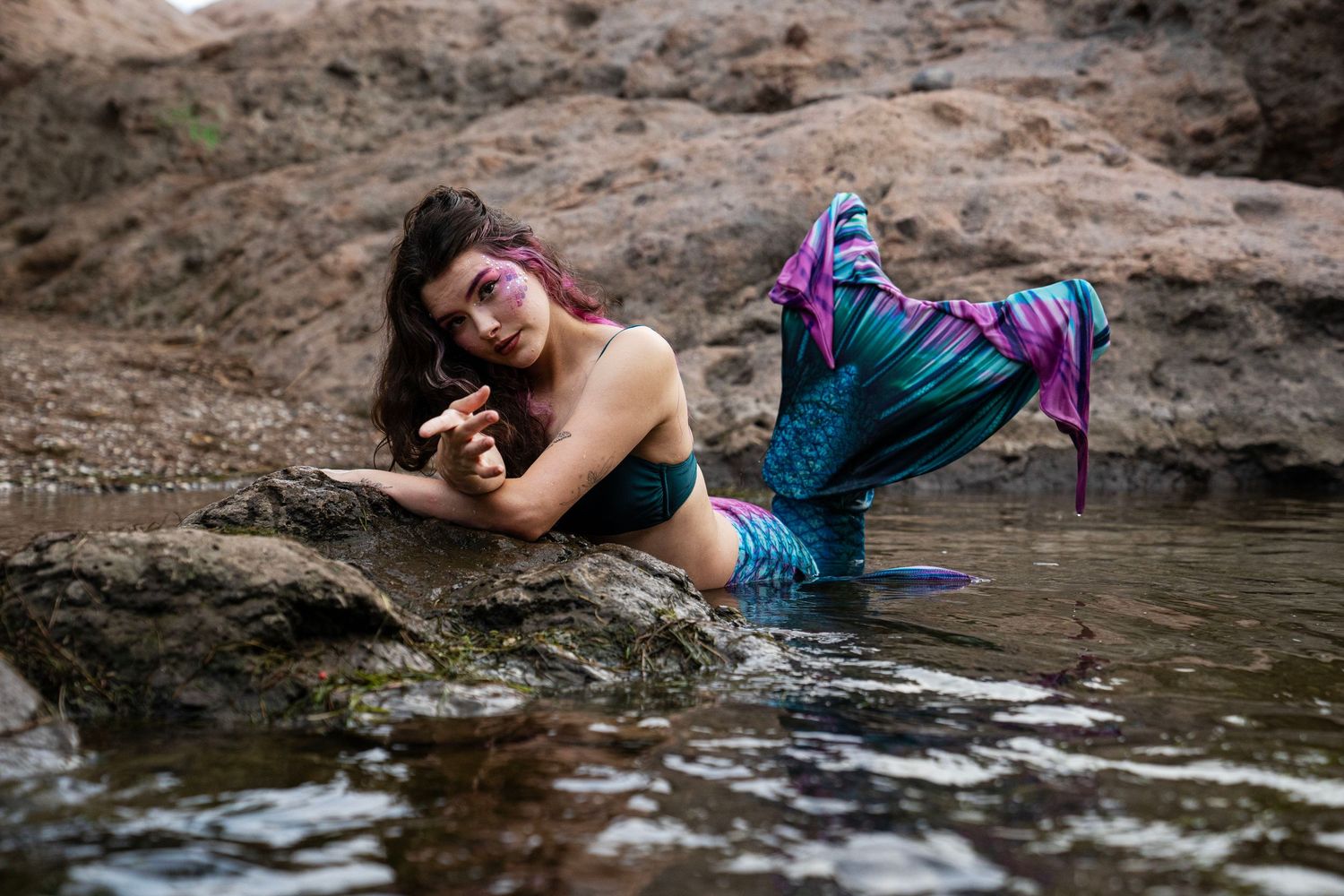 A woman dressed as a mermaid with a colorful tail lies on rocks in a shallow rocky stream. Fantasy Photoshoot