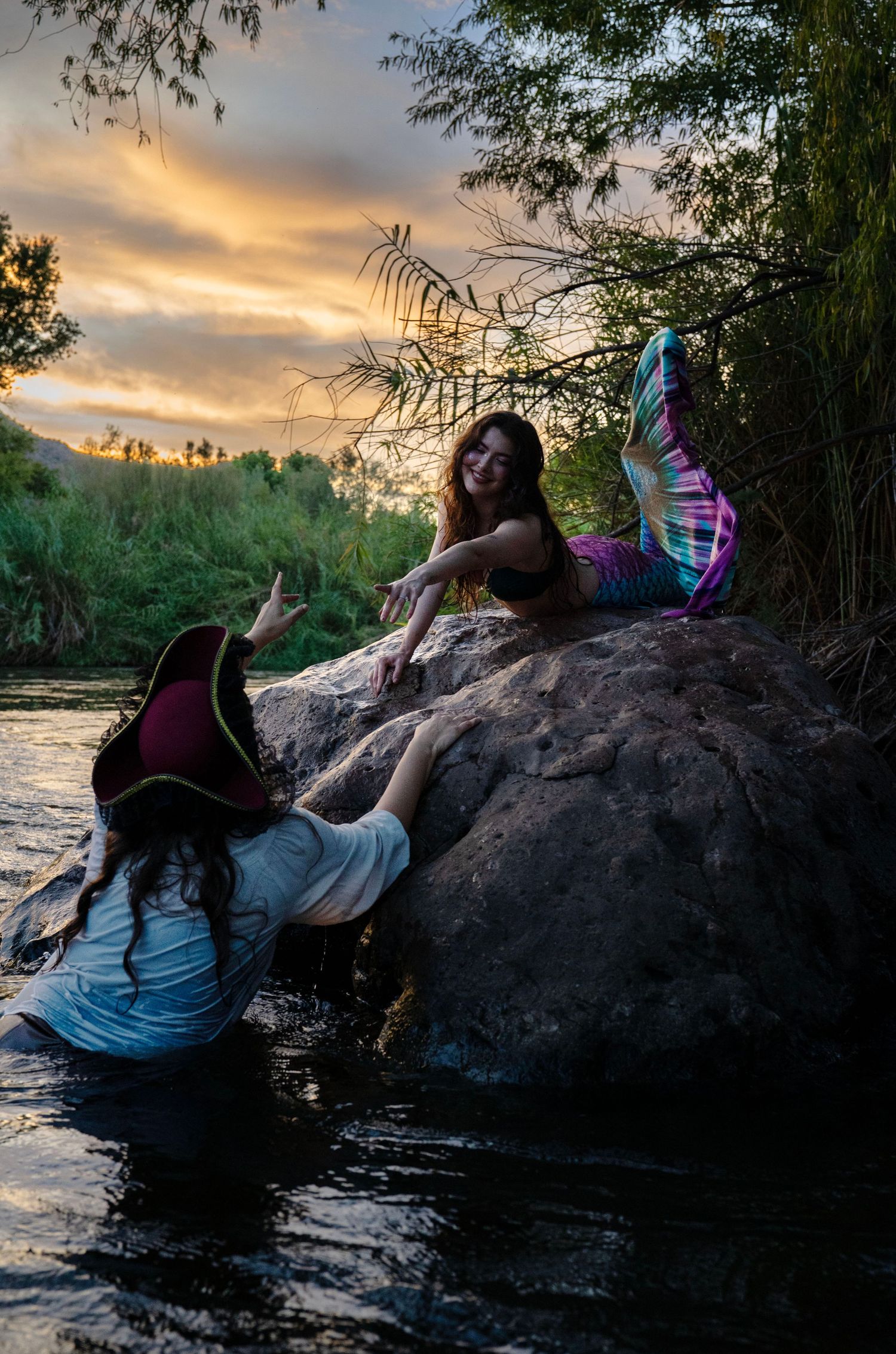 Pirate woman in water reaches up to mermaid on rock during dramatic sunset. Mermaid photoshoot, fantasy photoshoot. Arizona photographer.