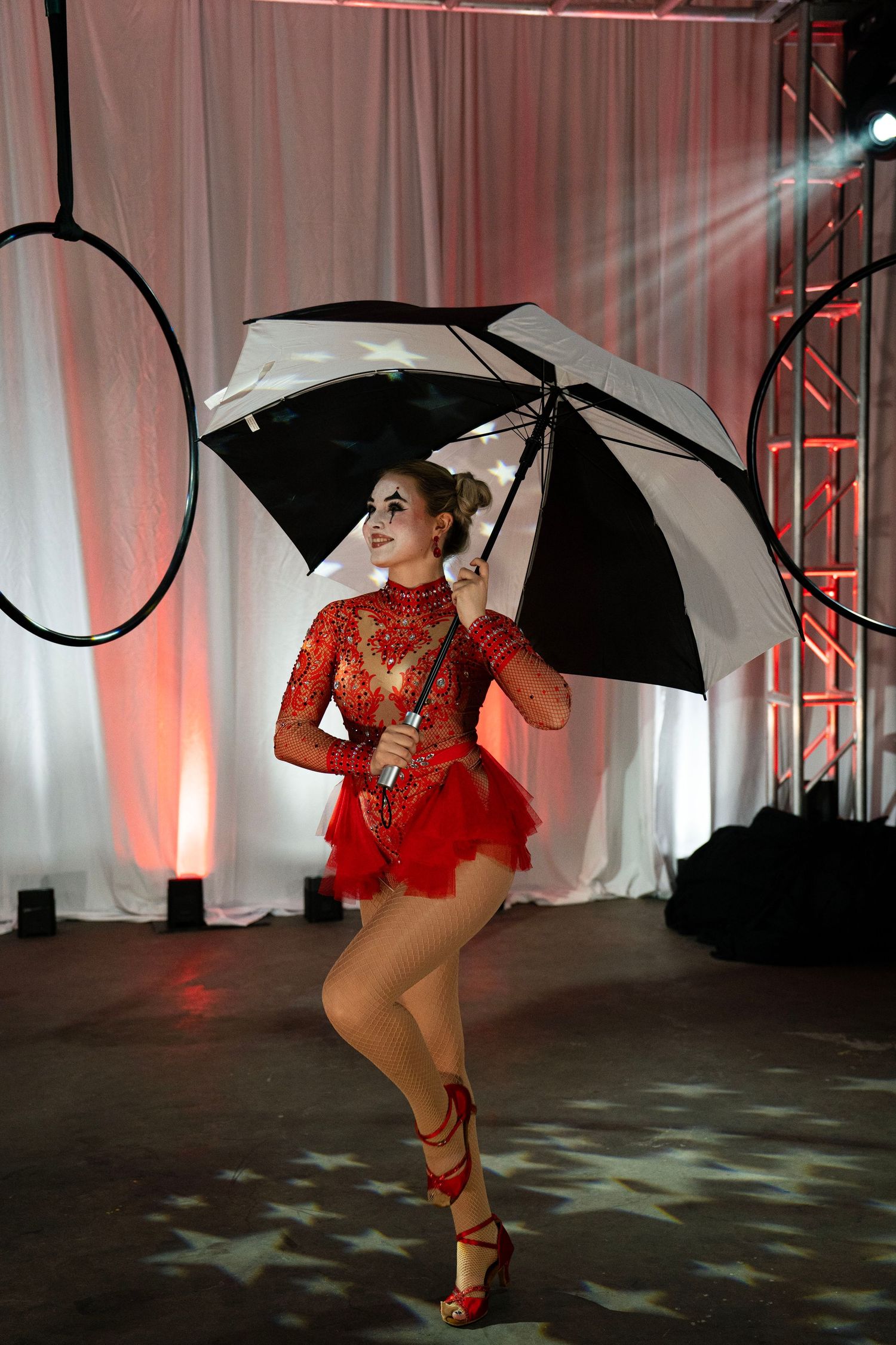A circus performer in red sequined costume with clown makeup poses with a black and white umbrella.