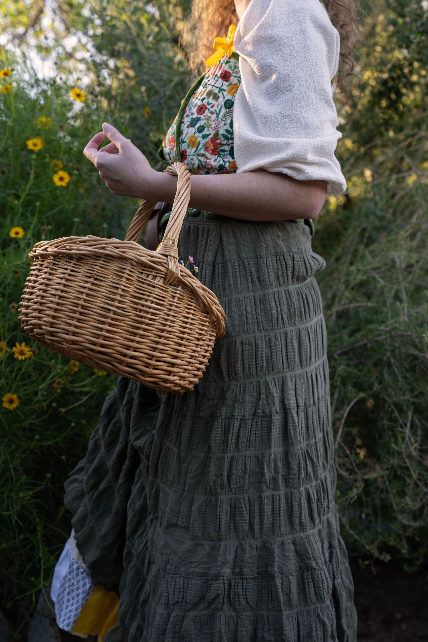 Woman in a green tiered skirt holds a wicker basket while standing beside yellow wildflowers in a sunlit garden.