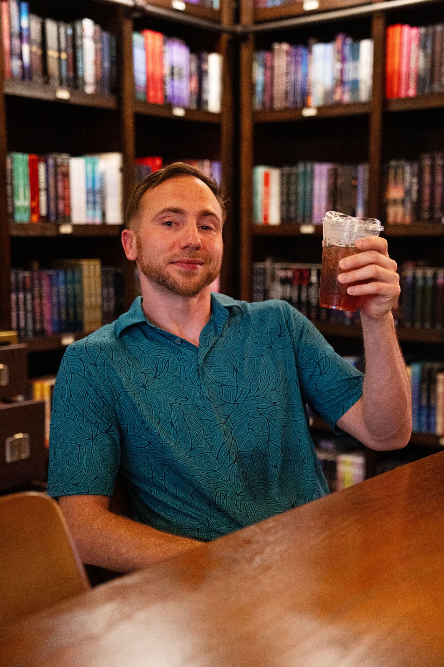 Man in teal shirt raising a glass of iced drink, smiling in a cozy library setting with colorful bookshelves.