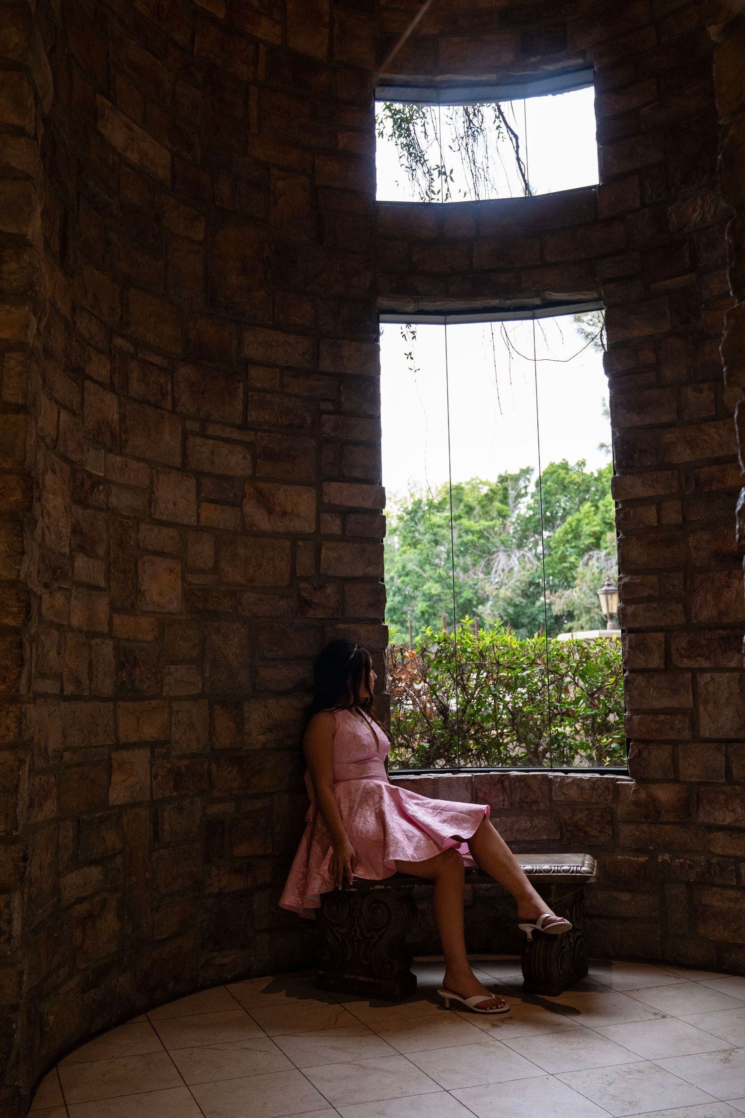 Woman in pink dress sits on stone bench gazing through arched window in rustic brick room.