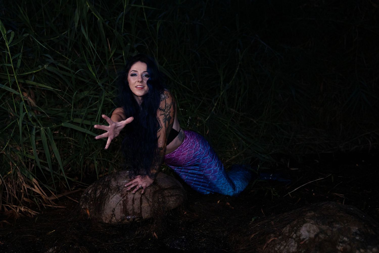 Woman in mermaid costume with colorful tail poses on rocks at night near tall grass.