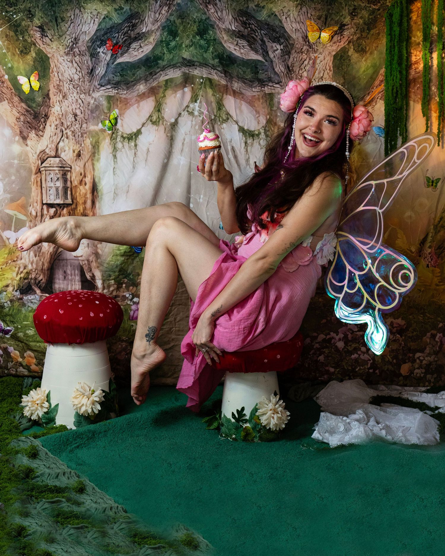 Laughing woman in pink fairy costume with iridescent wings sits on red mushroom holding cupcake in fantasy forest. Fantasy Photoshoot