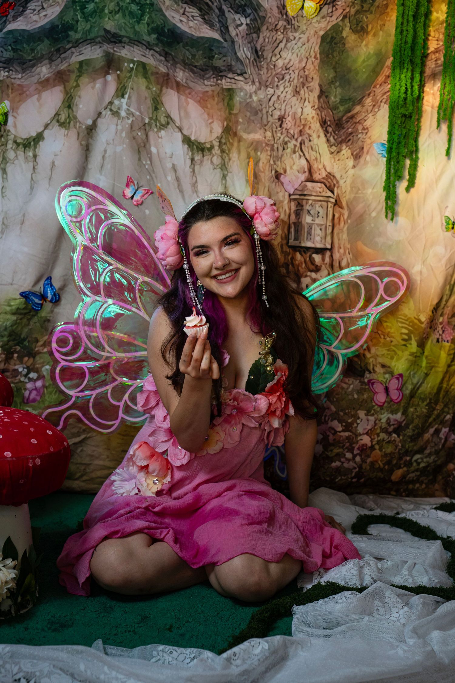 Woman in pink fairy costume with LED wings holds a cupcake in an enchanted forest studio setting. Fantasy Photoshoot