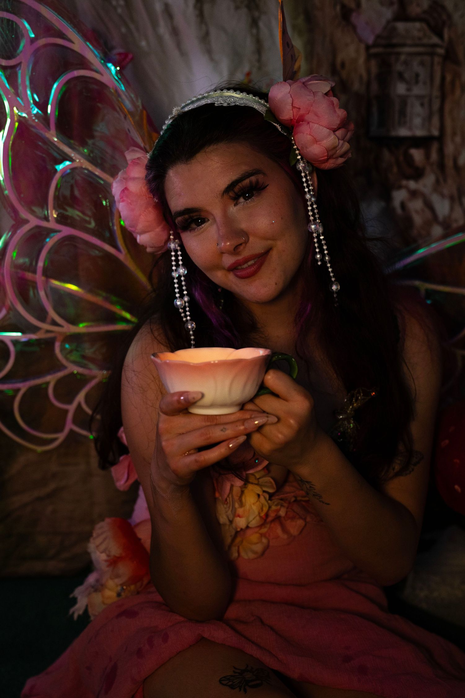 Young woman dressed as a fairy holds a glowing white cup, wearing pink flowers and iridescent wings. Fantasy Photoshoot