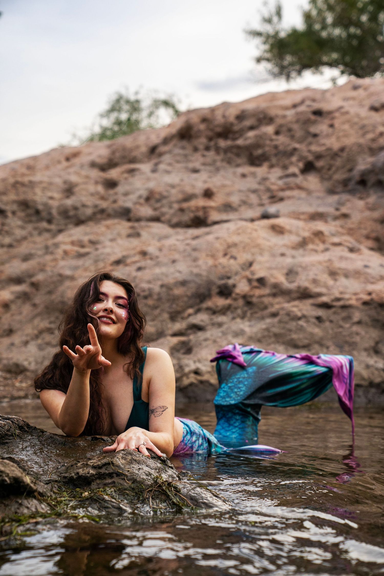 A smiling woman in a mermaid costume with colorful tail lies on rocks gesturing toward camera. Fantasy Photoshoot