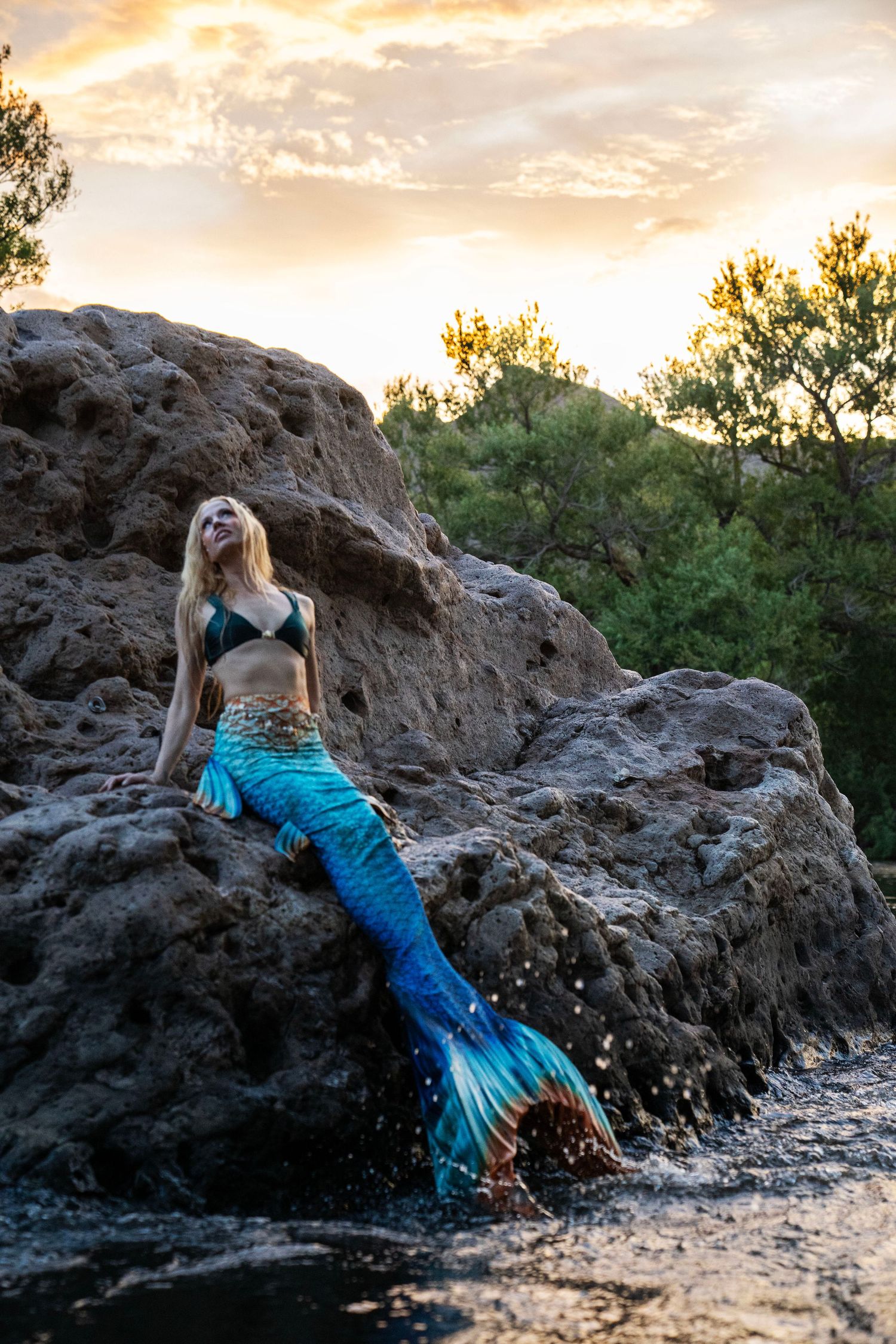Blonde woman in mermaid costume with blue tail resting on rocks by water at sunset. Mermaid photoshoot, fantasy photoshoot.