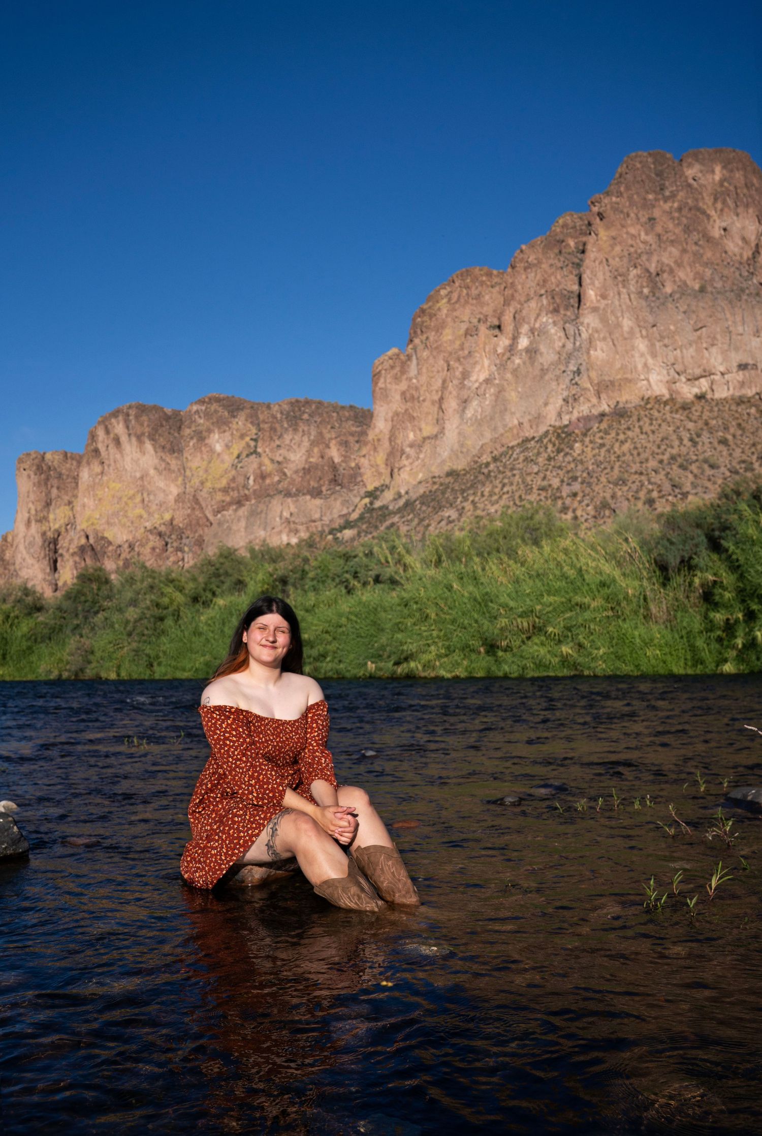 Woman in red floral dress sitting in a shallow river with towering rocky cliffs and green hills behind her.