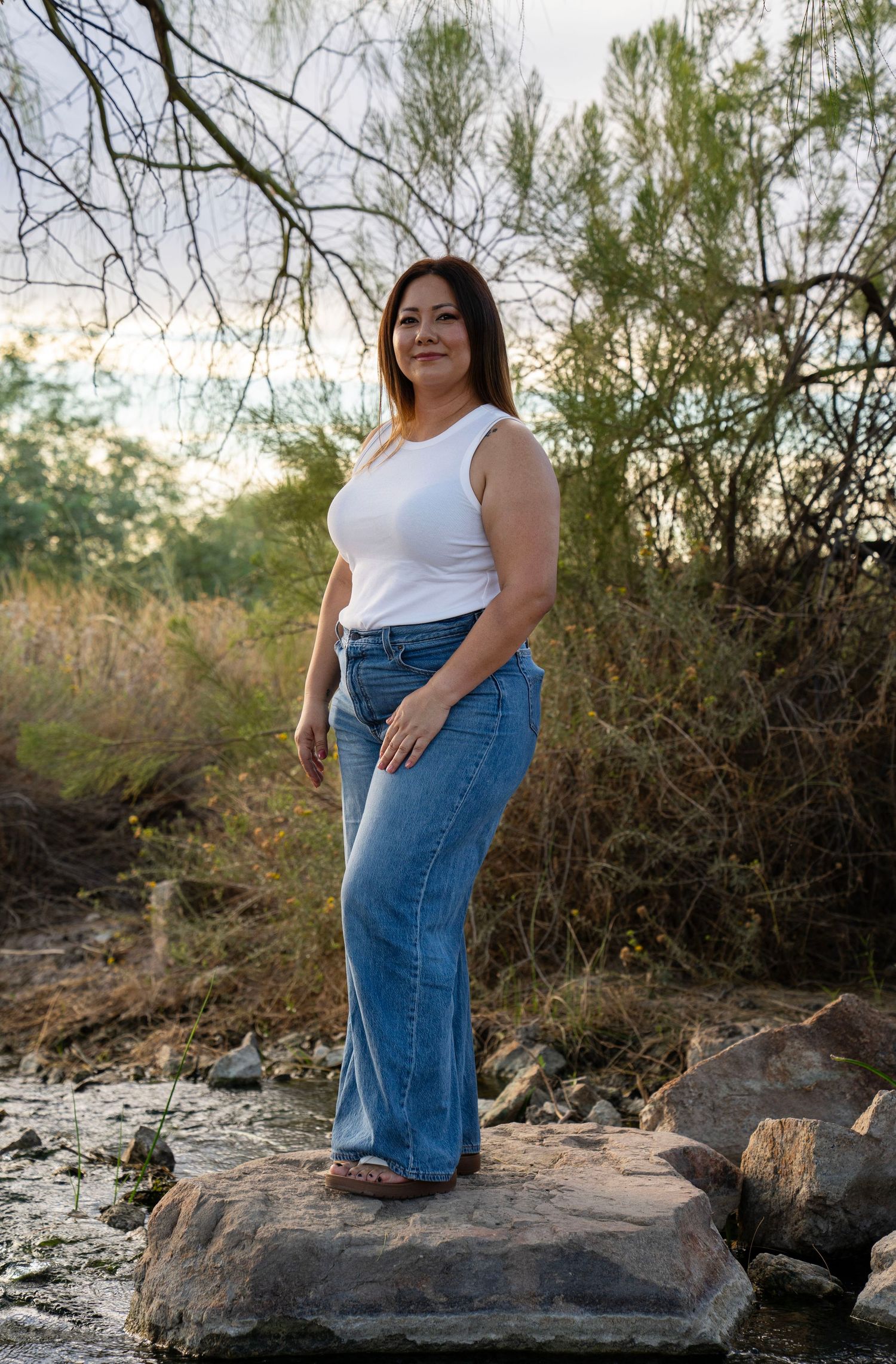 Woman in white tank top and flare jeans stands confidently on rocks near a peaceful stream in a natural setting.