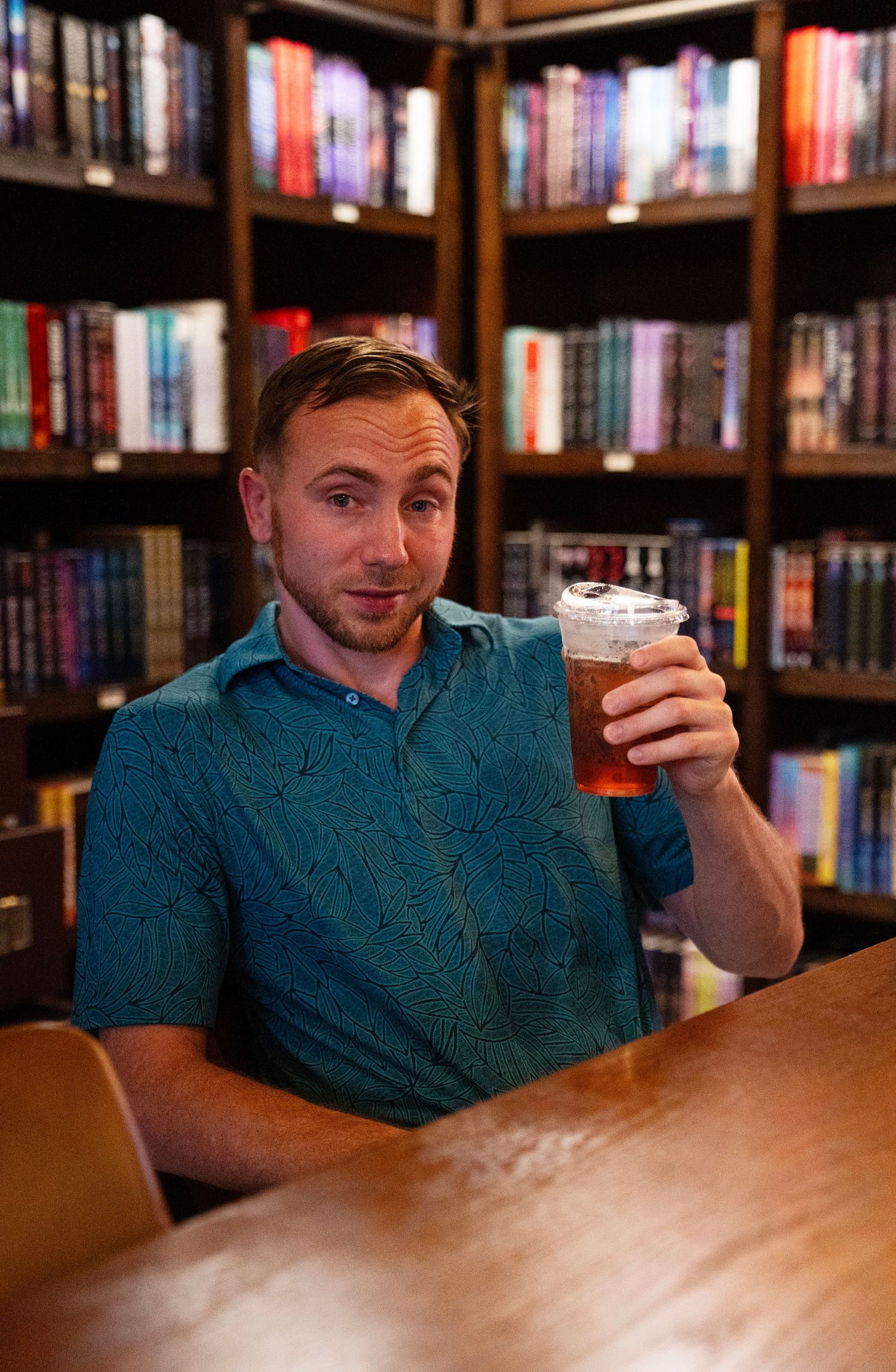 Man in teal shirt raising a pint of beer in a cozy library bar with colorful bookshelves.