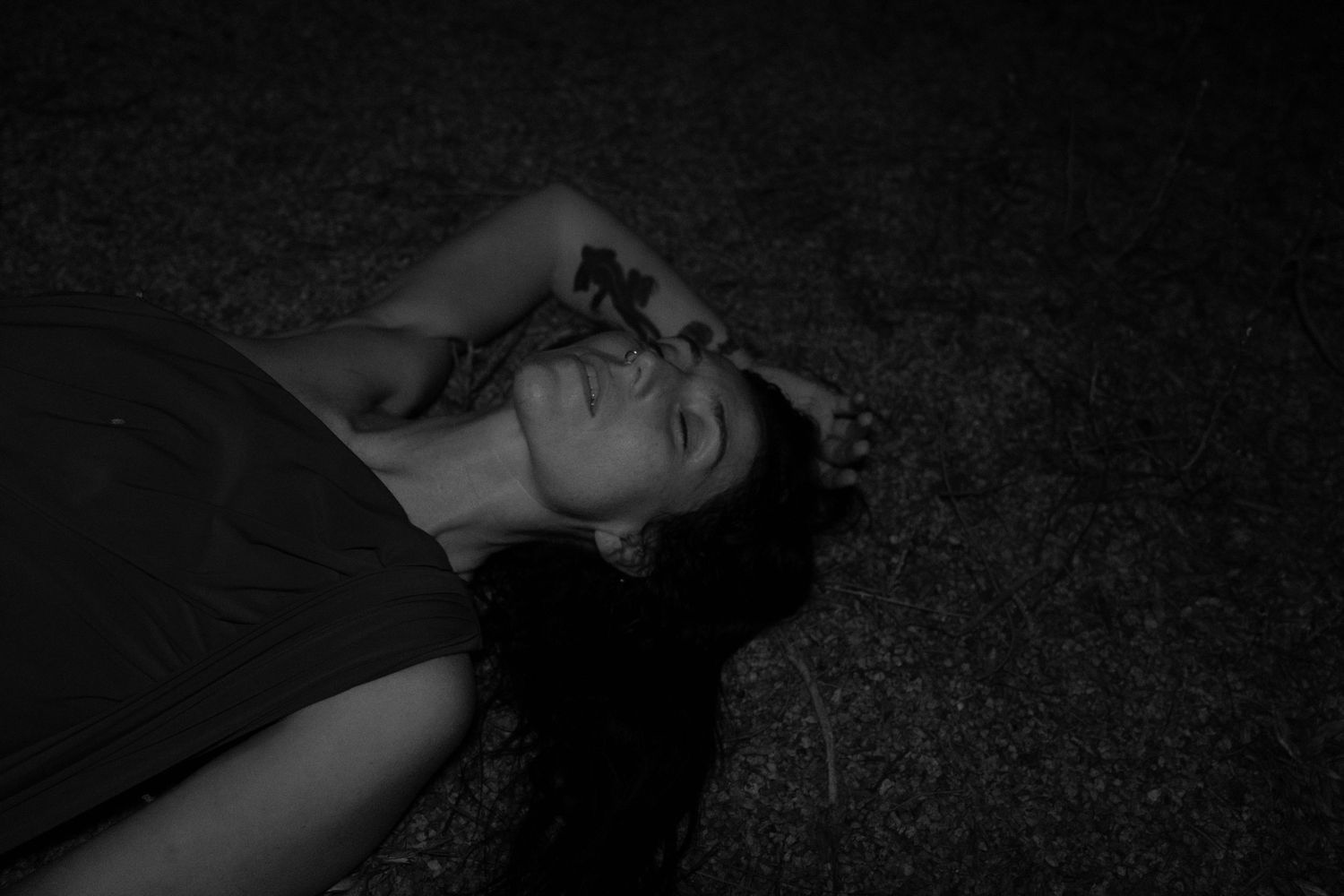 Black and white photo of a woman lying on her back on grass at night, one arm raised over her head as she rests with eyes closed.