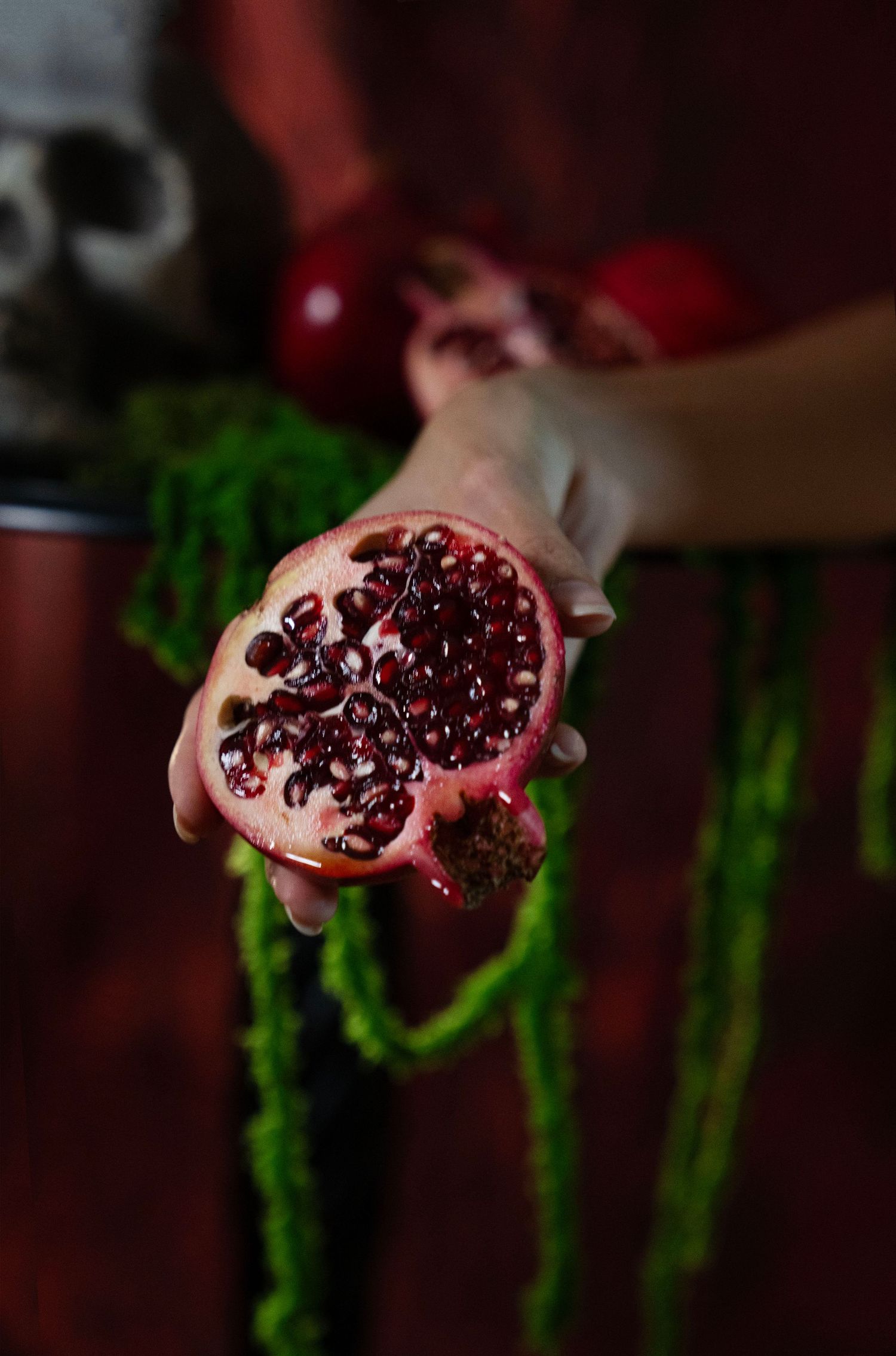 A hand holds a halved pomegranate with glistening red seeds in front of dangling green moss and a blurred skull in a moody setting.