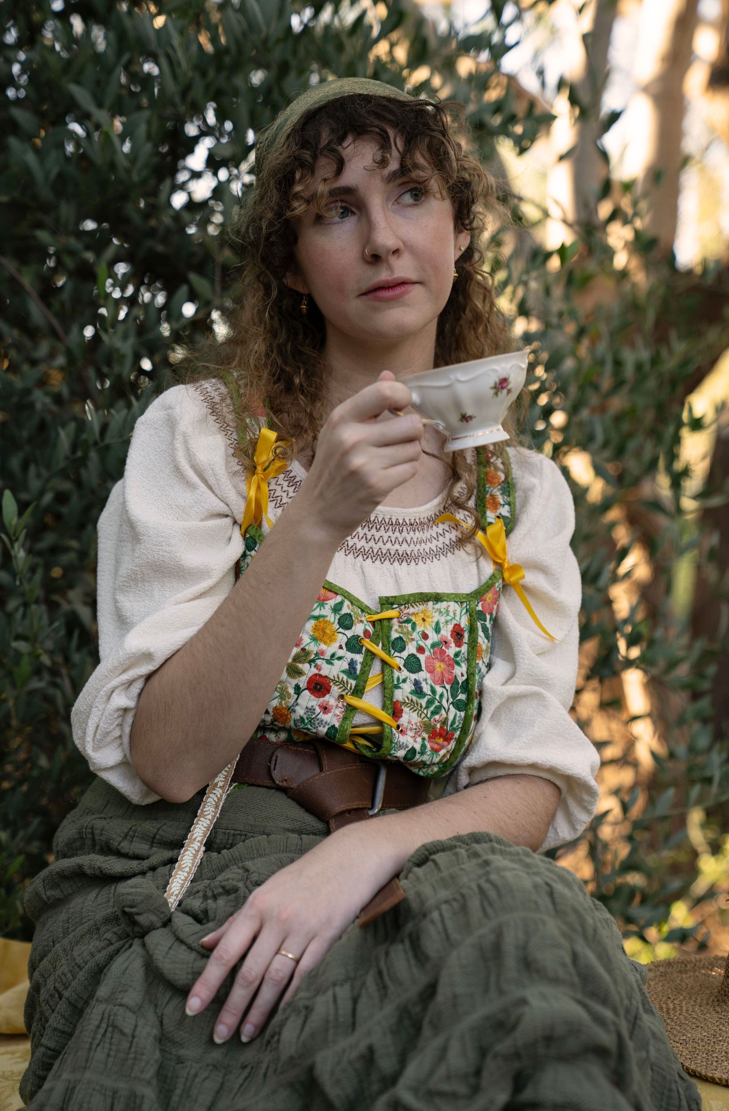 Woman with curly hair sips tea from a floral teacup, wearing a cottagecore outfit with a floral vest and green skirt outdoors.