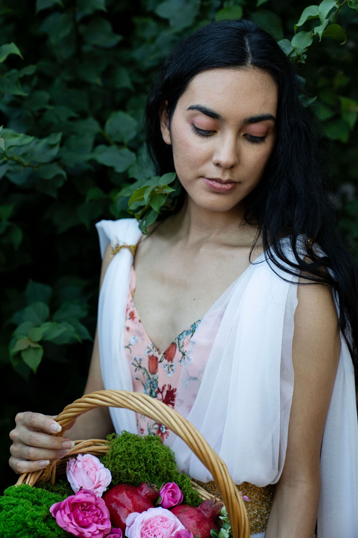 Woman with long dark hair in a sheer white and floral dress looks down at a woven basket of pink roses and moss in a lush green garden.