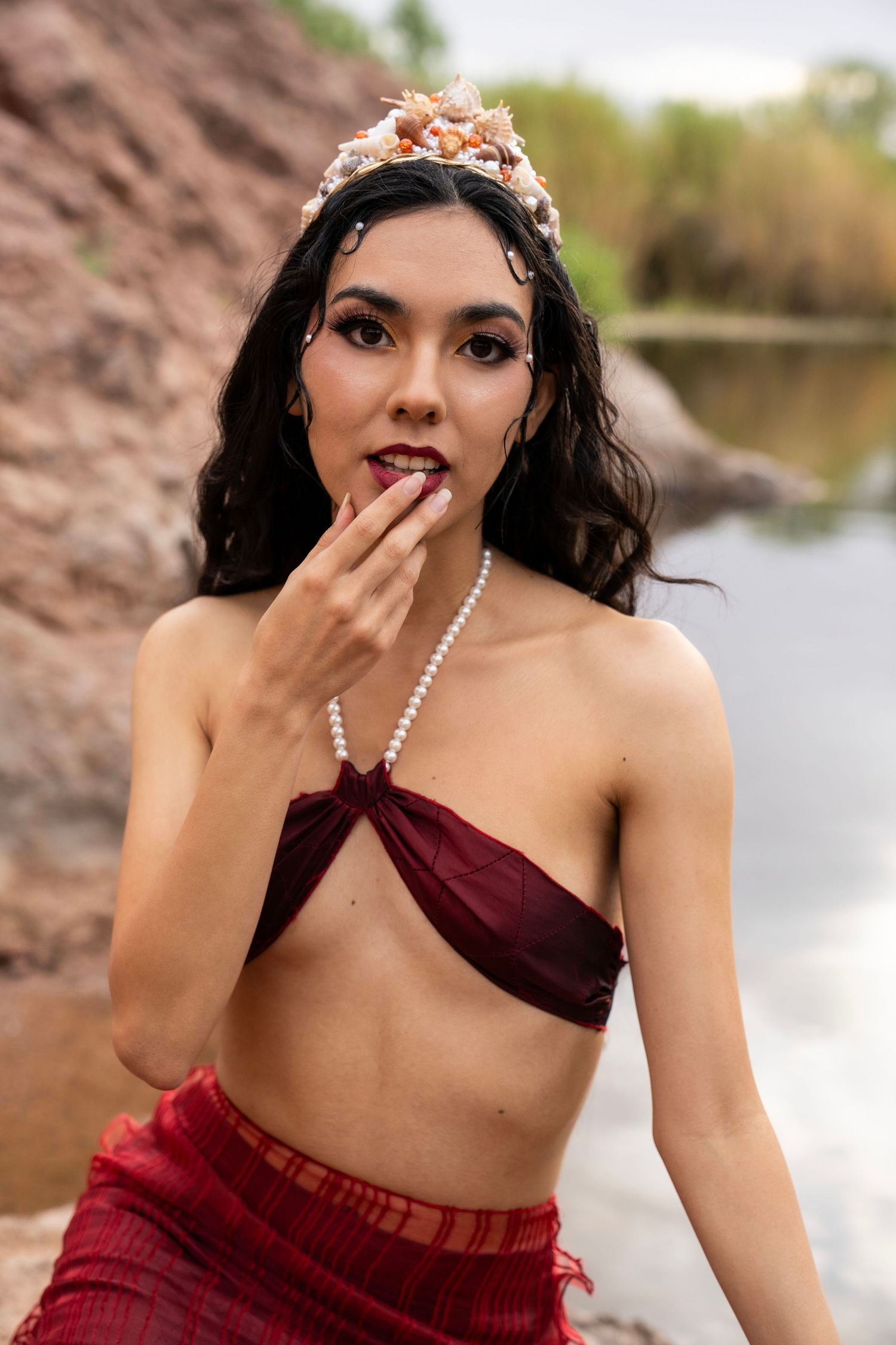 A woman dressed as a mermaid with a shell crown and pearl necklace touches her lips while sitting near a river.