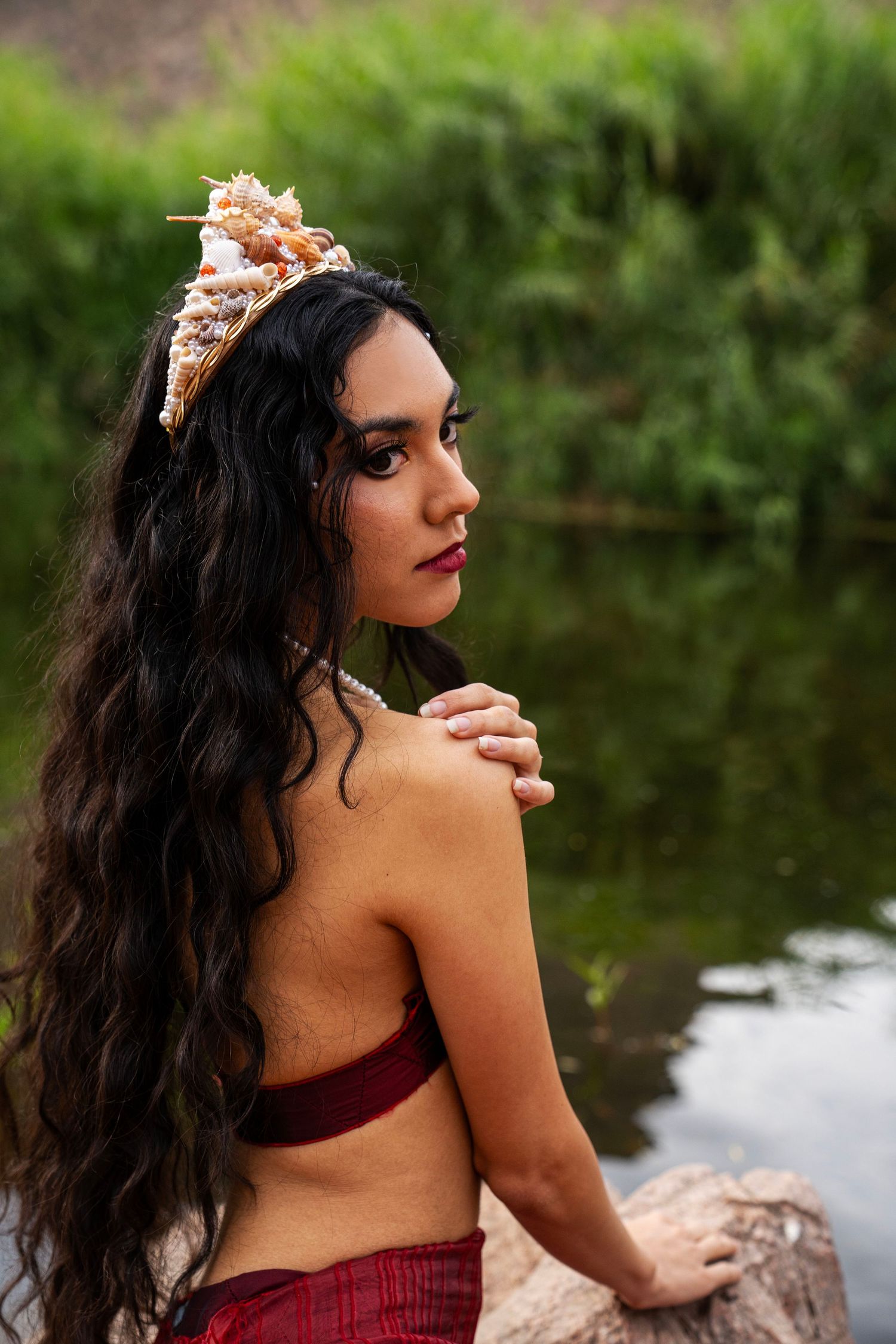 Woman with long dark hair wearing a seashell crown and red top sits by a green riverside.