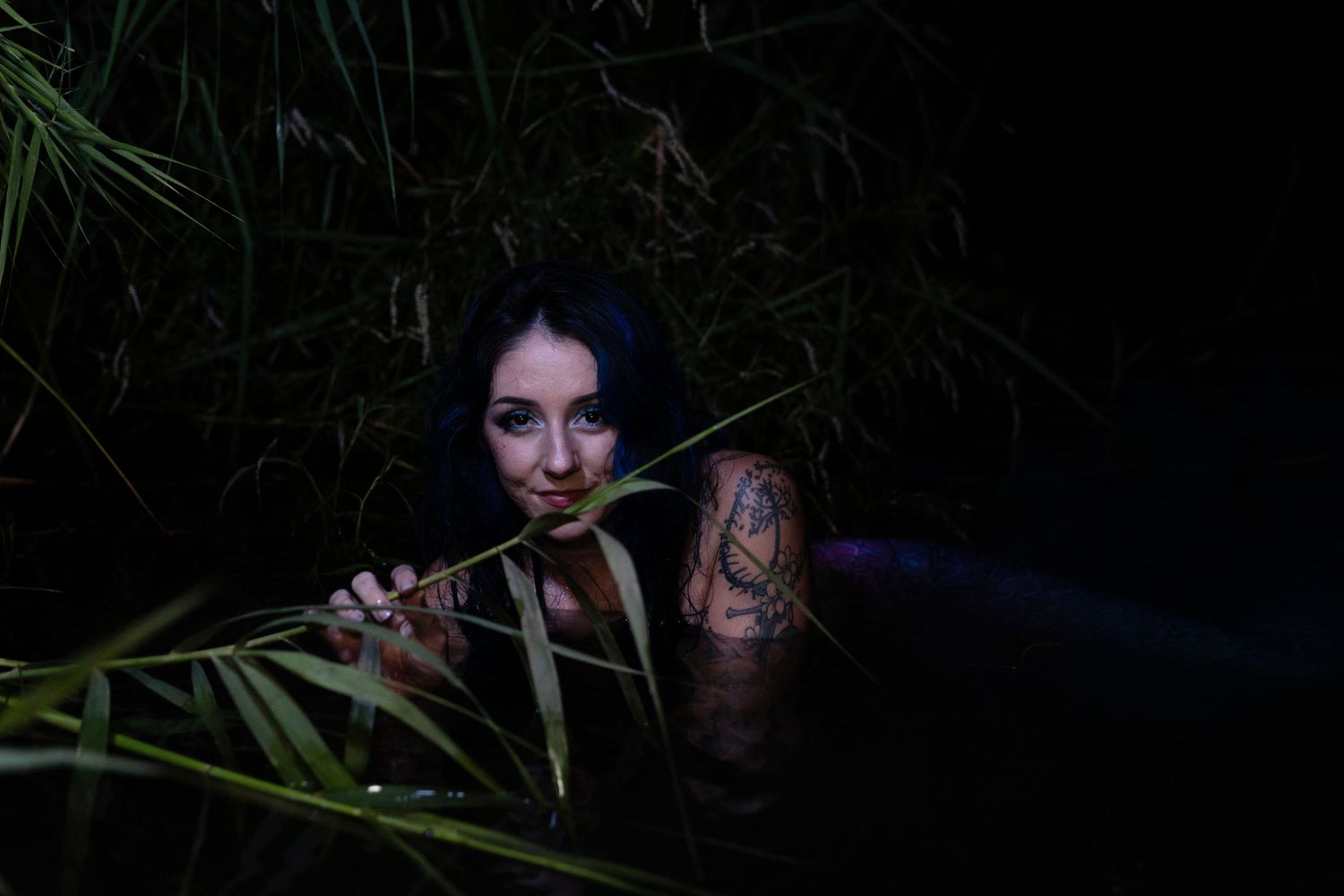 Woman with tattoos partially submerged in dark water surrounded by reeds at night.
