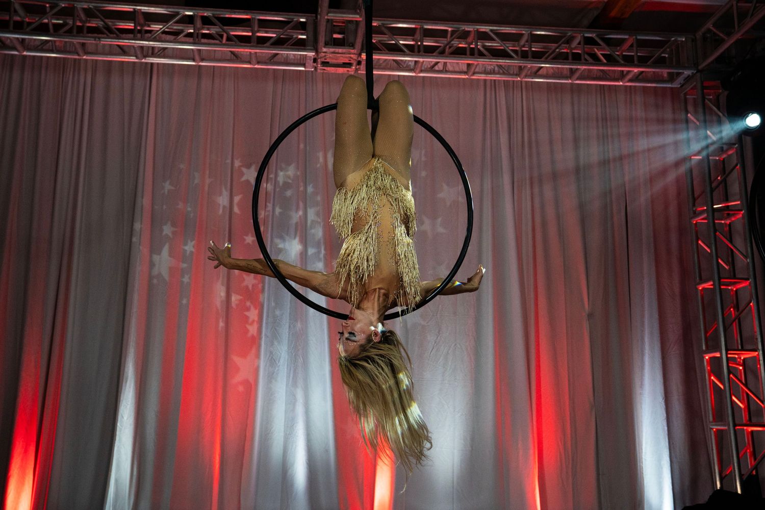 Blonde acrobat performing inverted pose on aerial lyra hoop at indoor circus show.