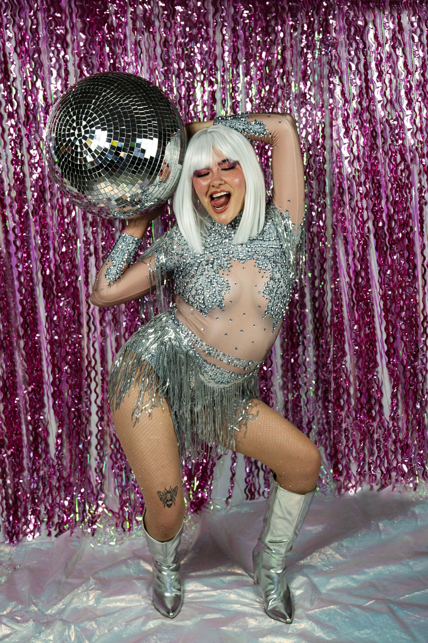 Energetic performer in silver rhinestone fringe costume holds disco ball overhead, wearing white wig and silver boots.