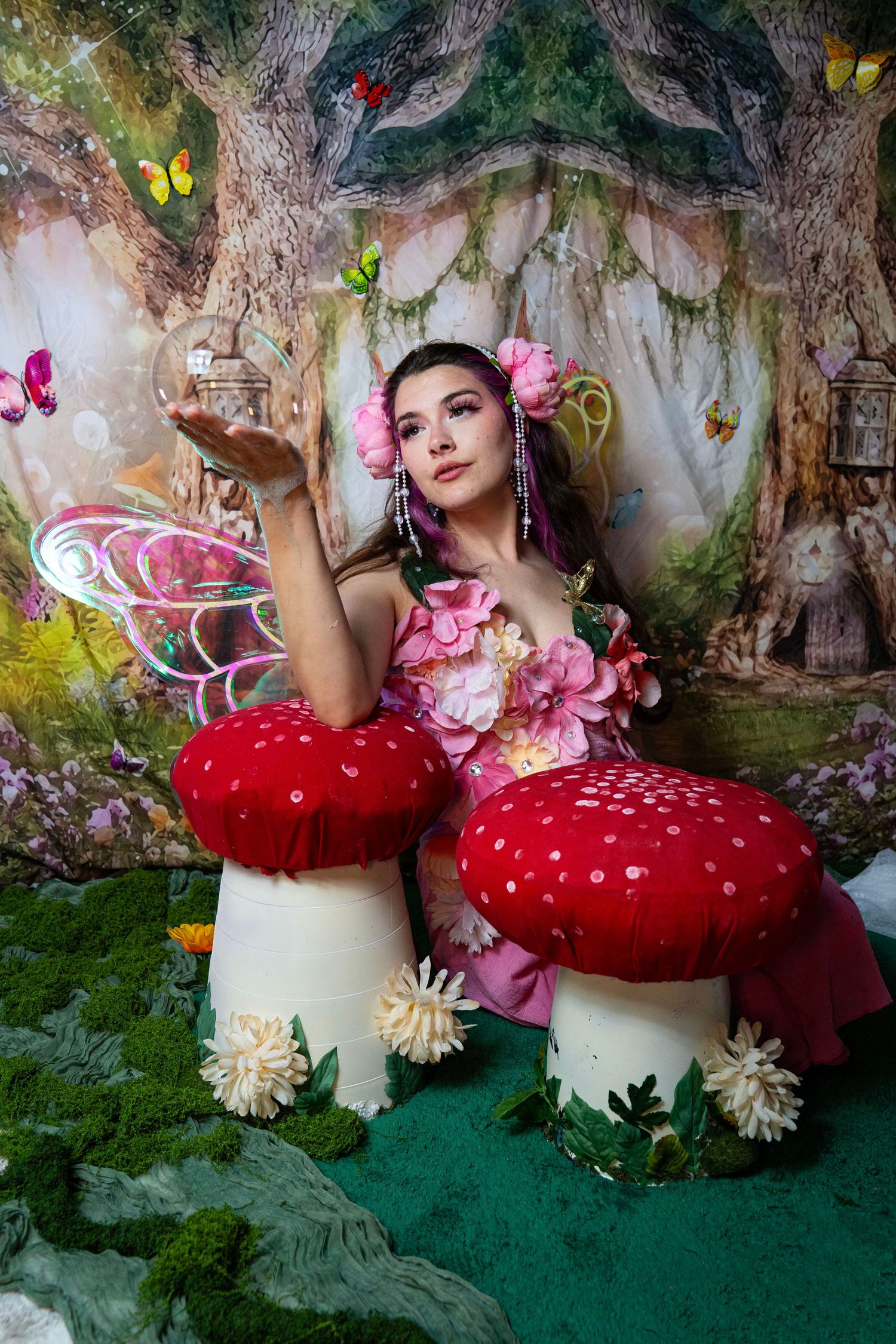 Fairy costume model in pink floral dress reclines on giant red mushrooms in magical enchanted forest studio. Fantasy Photoshoot