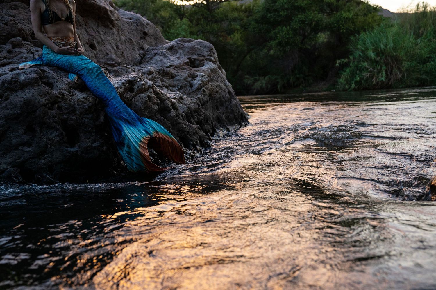 A colorful mermaid tail costume draped over rocks beside a flowing river at golden sunset. Mermaid photoshoot, fantasy photoshoot.