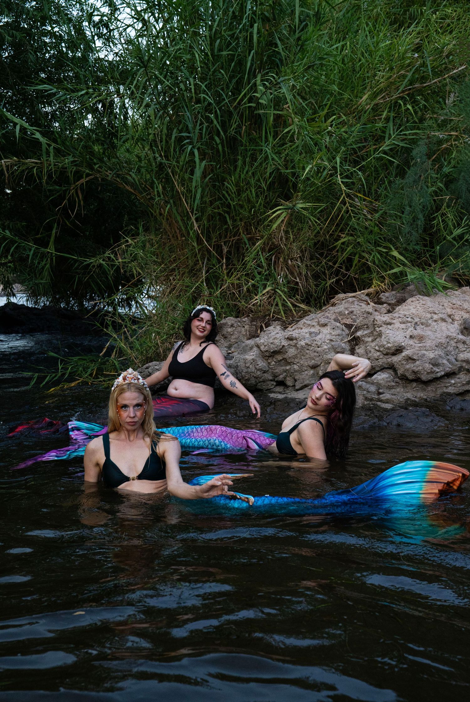 Three women in colorful mermaid tails posing together in a natural river with lush greenery. Fantasy Photoshoot