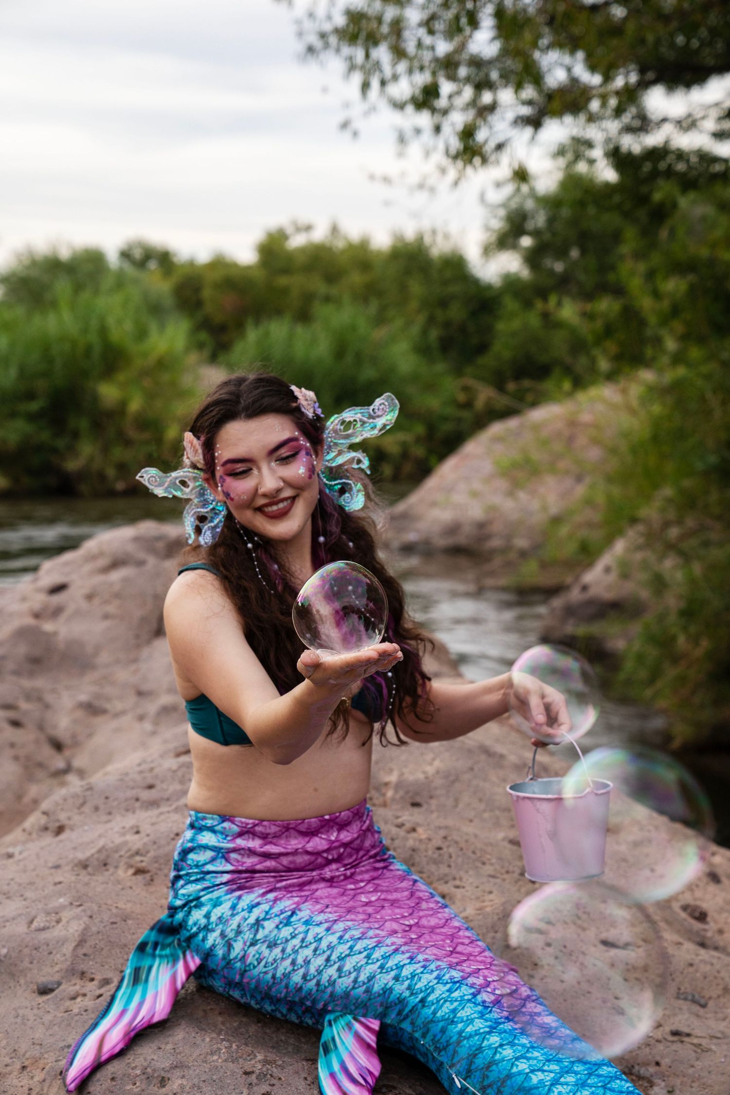 Smiling woman in mermaid costume holding a large soap bubble while sitting on rocks by a river. Fantasy Photoshoot