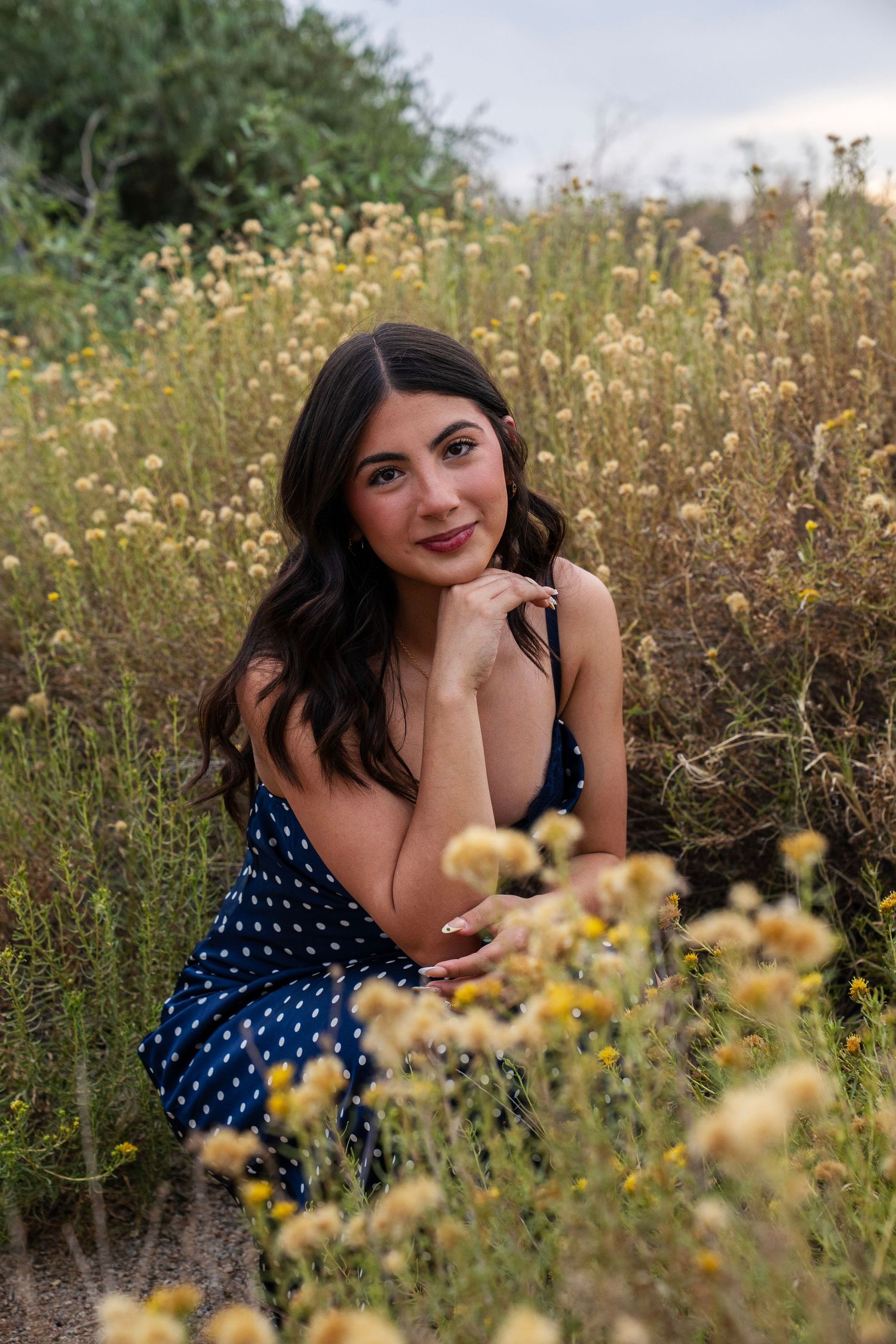 Young woman in navy polka dot dress sitting among yellow wildflowers.