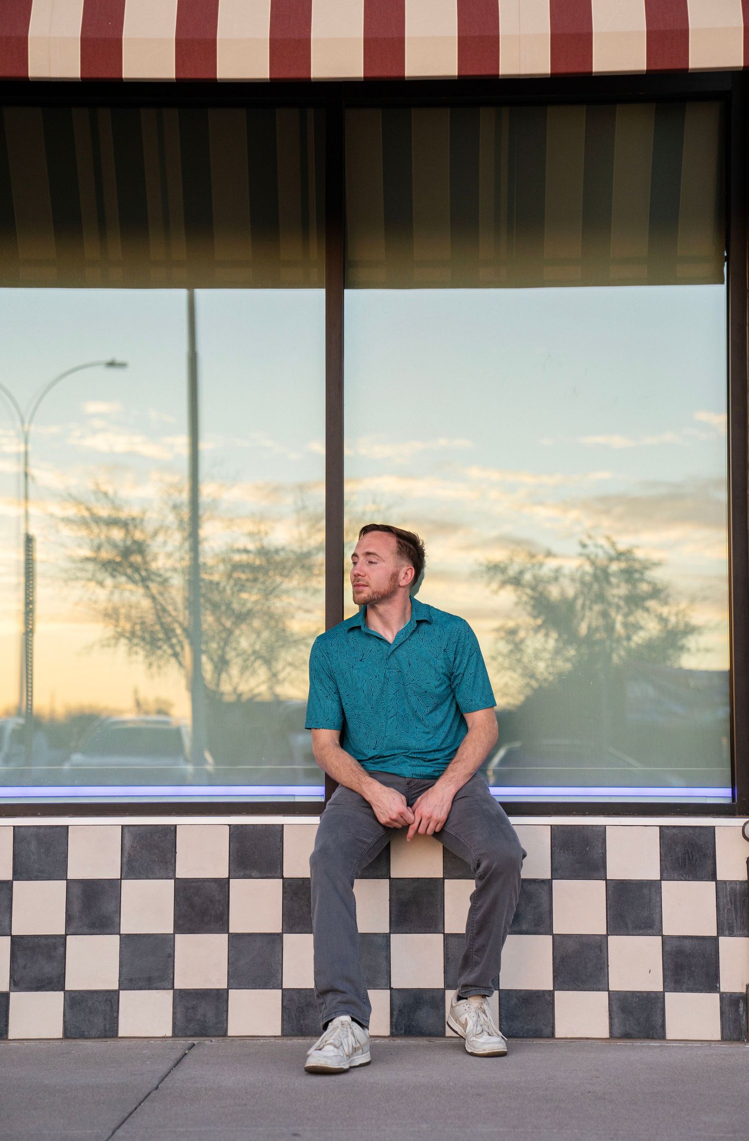 Man in teal polo sits on checkered ledge outside retro storefront at sunset.