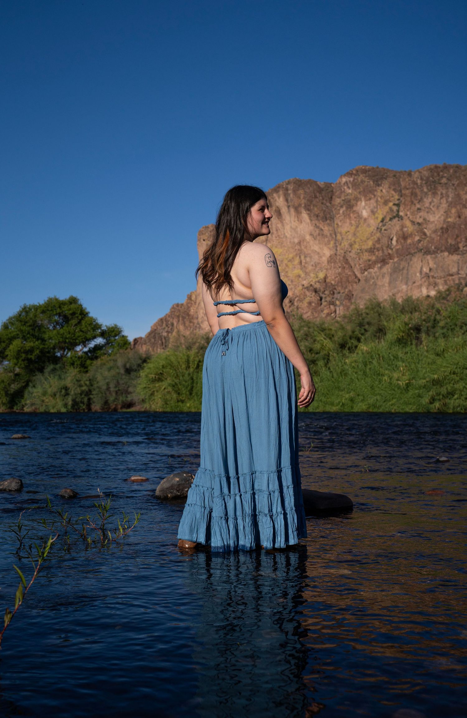 Woman in blue maxi skirt standing in river with rocky mountain and blue sky backdrop.