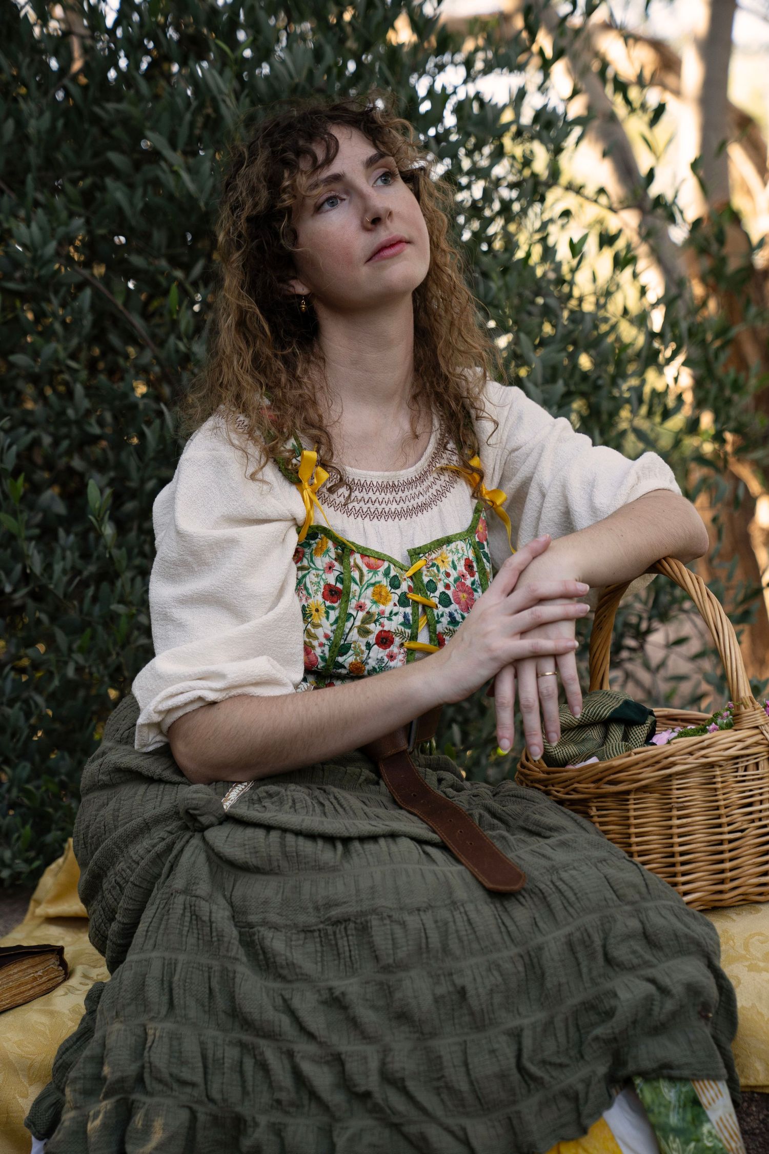 Woman with curly hair in a cream peasant blouse, floral bodice and olive green skirt sits beside a wicker basket in a sunlit garden.