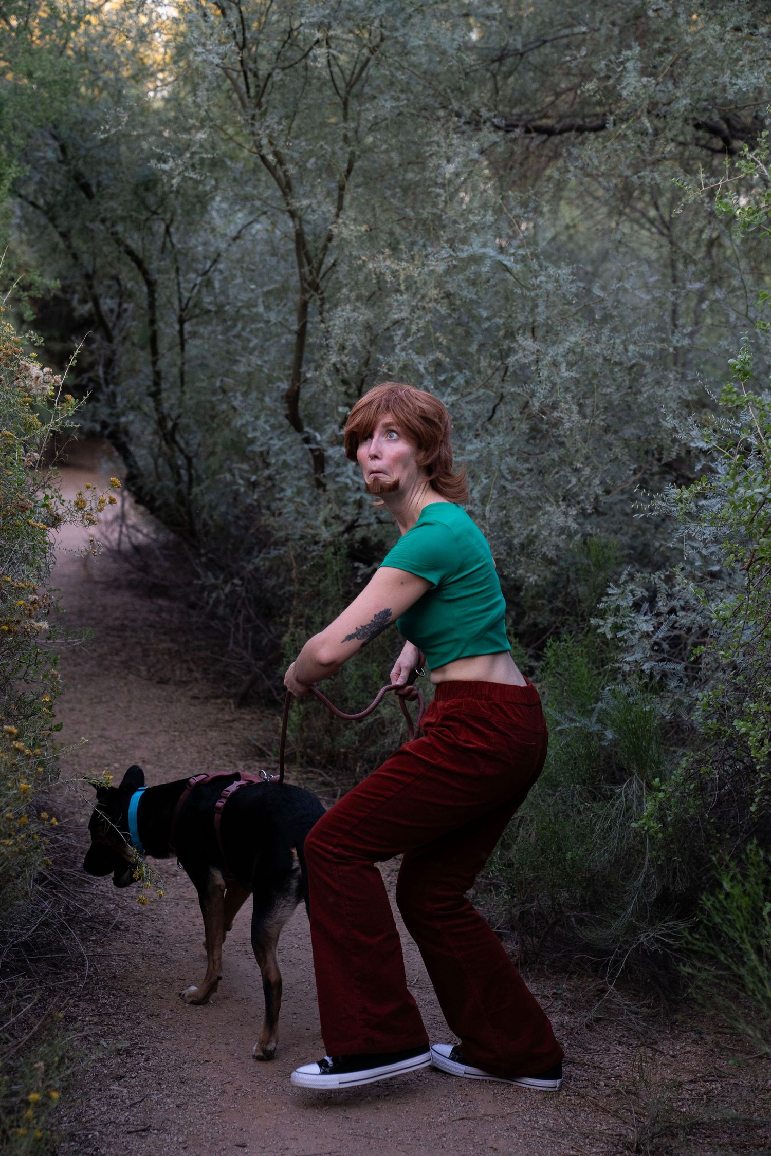 A young woman with red hair in a green crop top and red flared pants walks a dog on a forest trail.