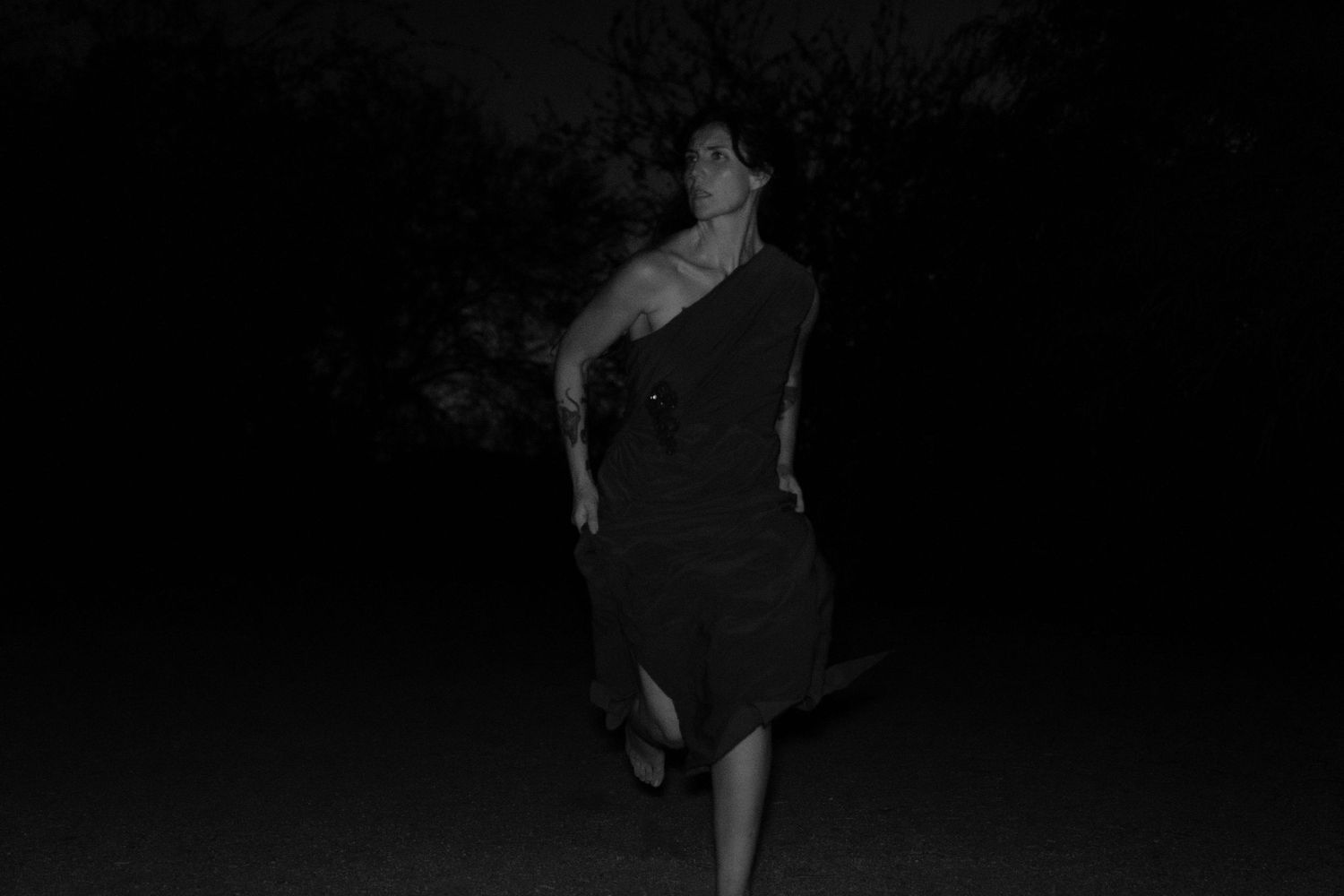 Black and white photo of a woman in a dark one-shoulder dress walking mid-stride through a shadowy outdoor setting at night.