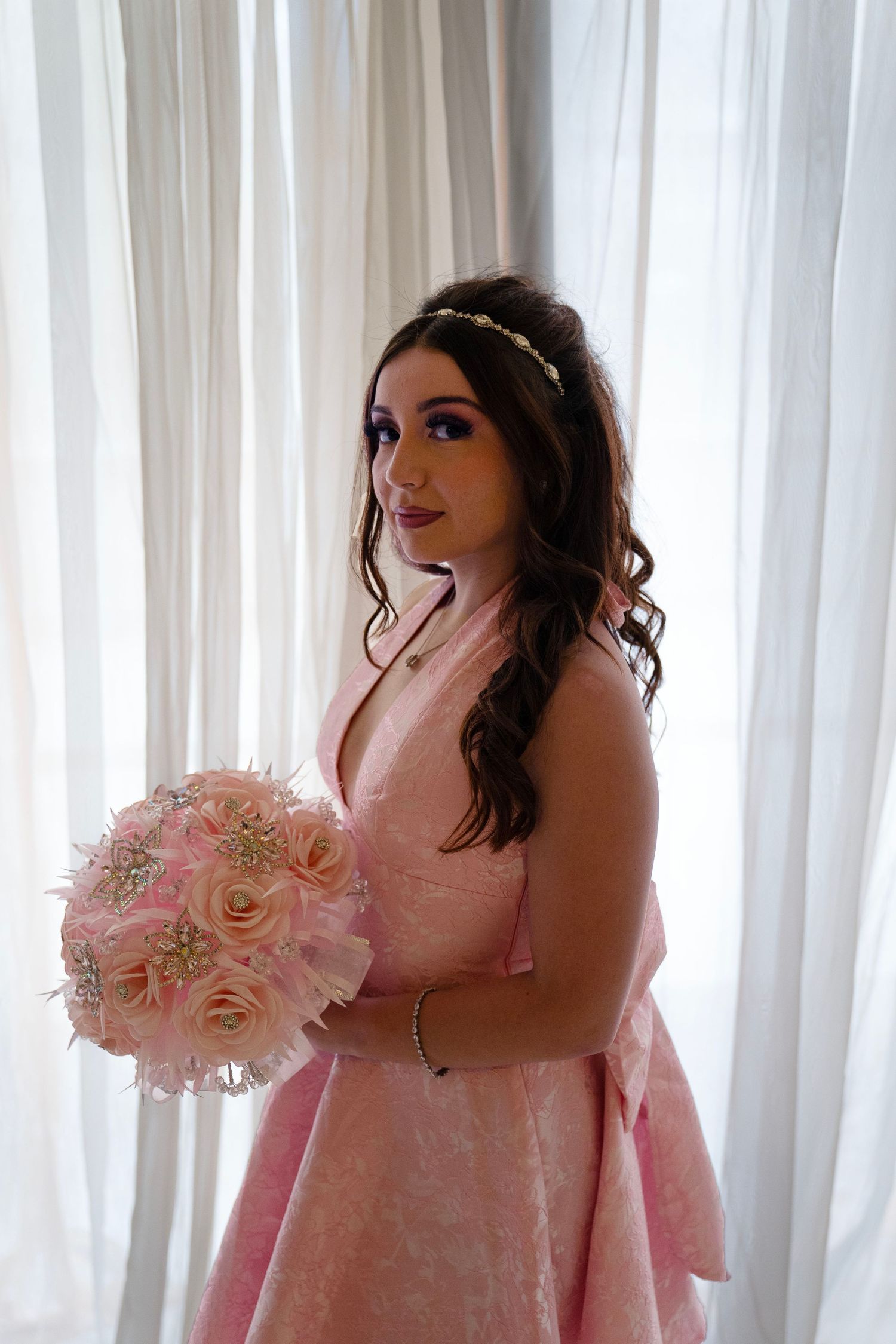 Young woman in pink dress holding floral bouquet standing by white curtains.