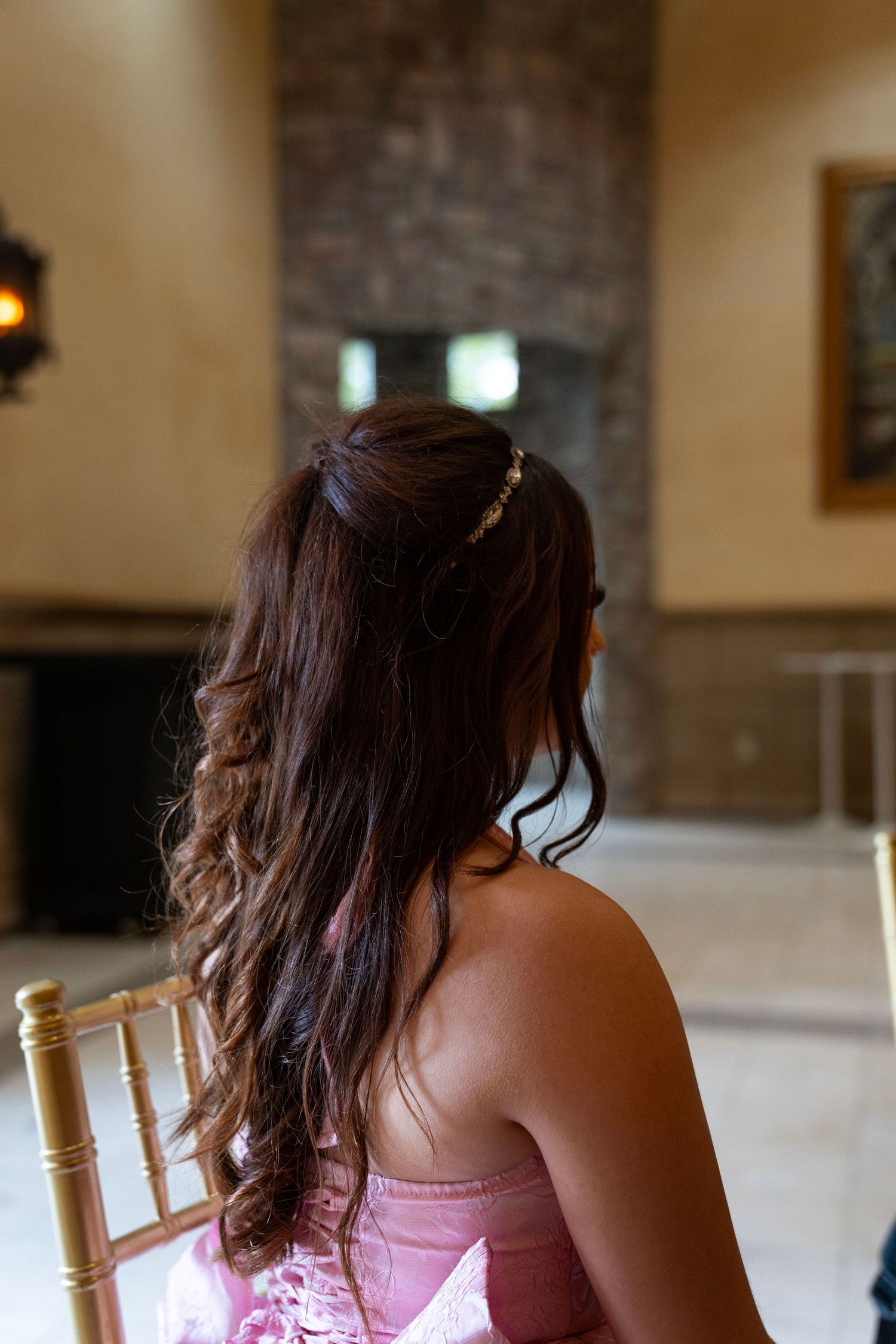 Back view of woman with long curly brown hair and rhinestone headband wearing a pink strapless dress indoors.