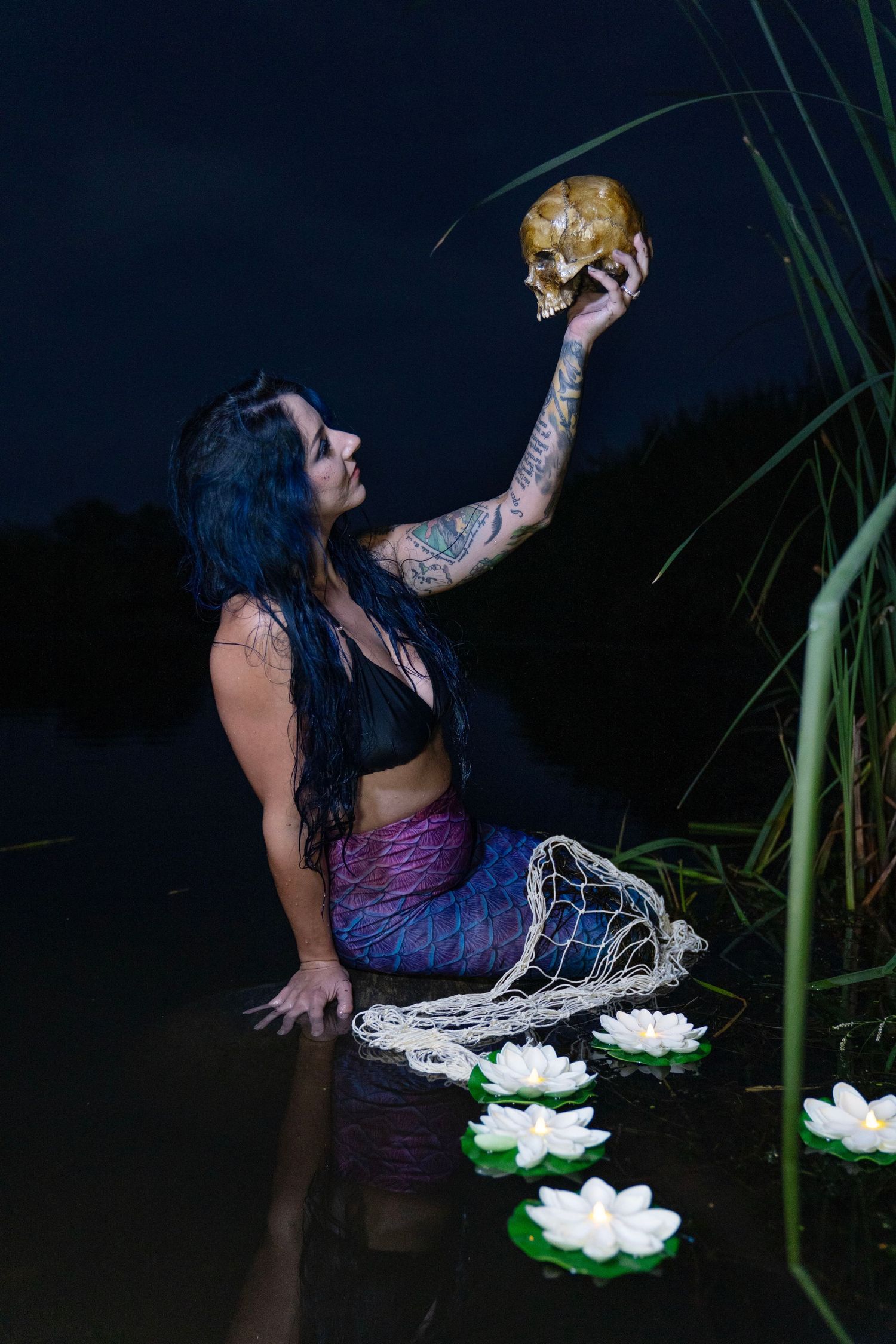 Tattooed woman in mermaid costume holding skull aloft in dark river with white lotus flowers.