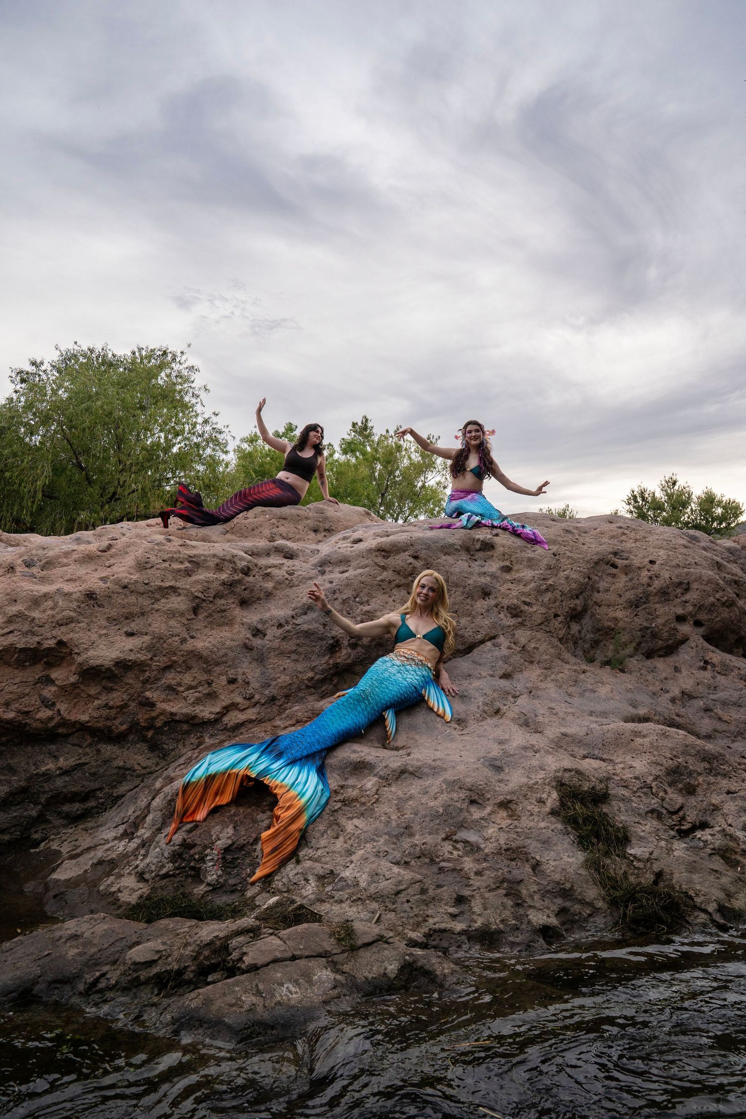 Three women wearing mermaid tails pose on rocky terrain near a river under a cloudy sky. Fantasy Photoshoot