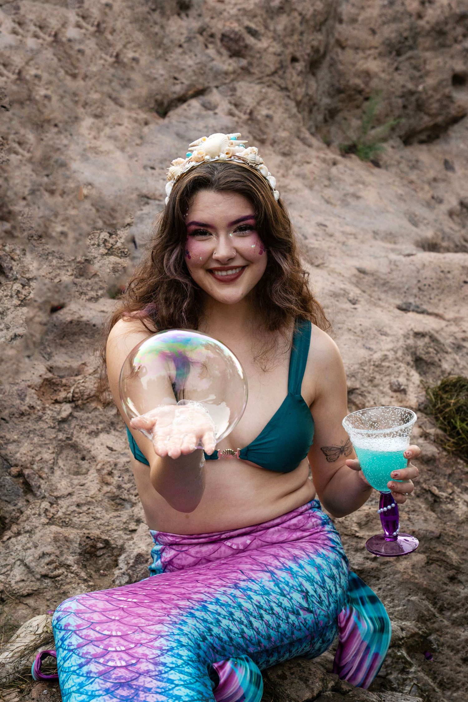 A smiling brunette mermaid with pink makeup holds a large soap bubble and blue cocktail on rocks. Fantasy Photoshoot