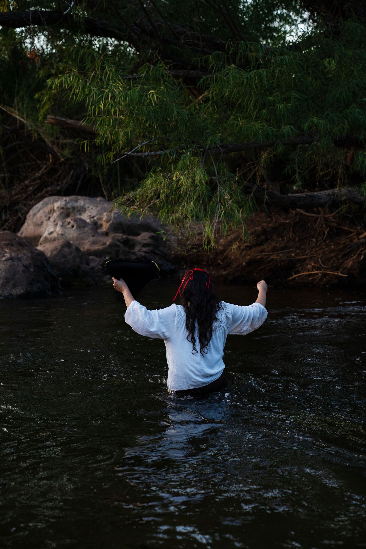 A woman in a white shirt raises her arms while standing waist-deep in a dark flowing river near green trees. Mermaid photoshoot, fantasy pho