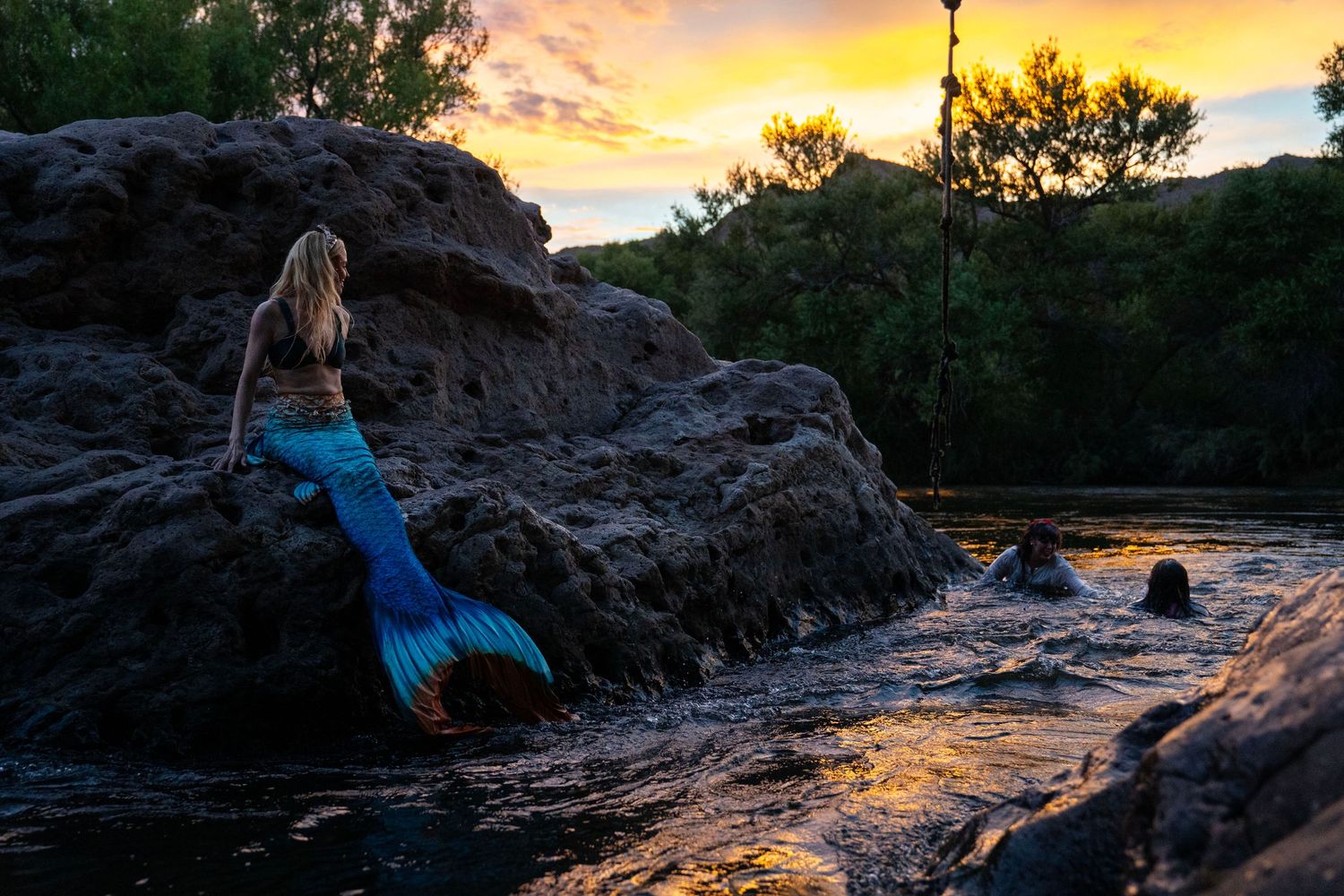 Woman in mermaid tail costume rests on rocks by river at sunset. Mermaid photoshoot, fantasy photoshoot. Arizona photographer.