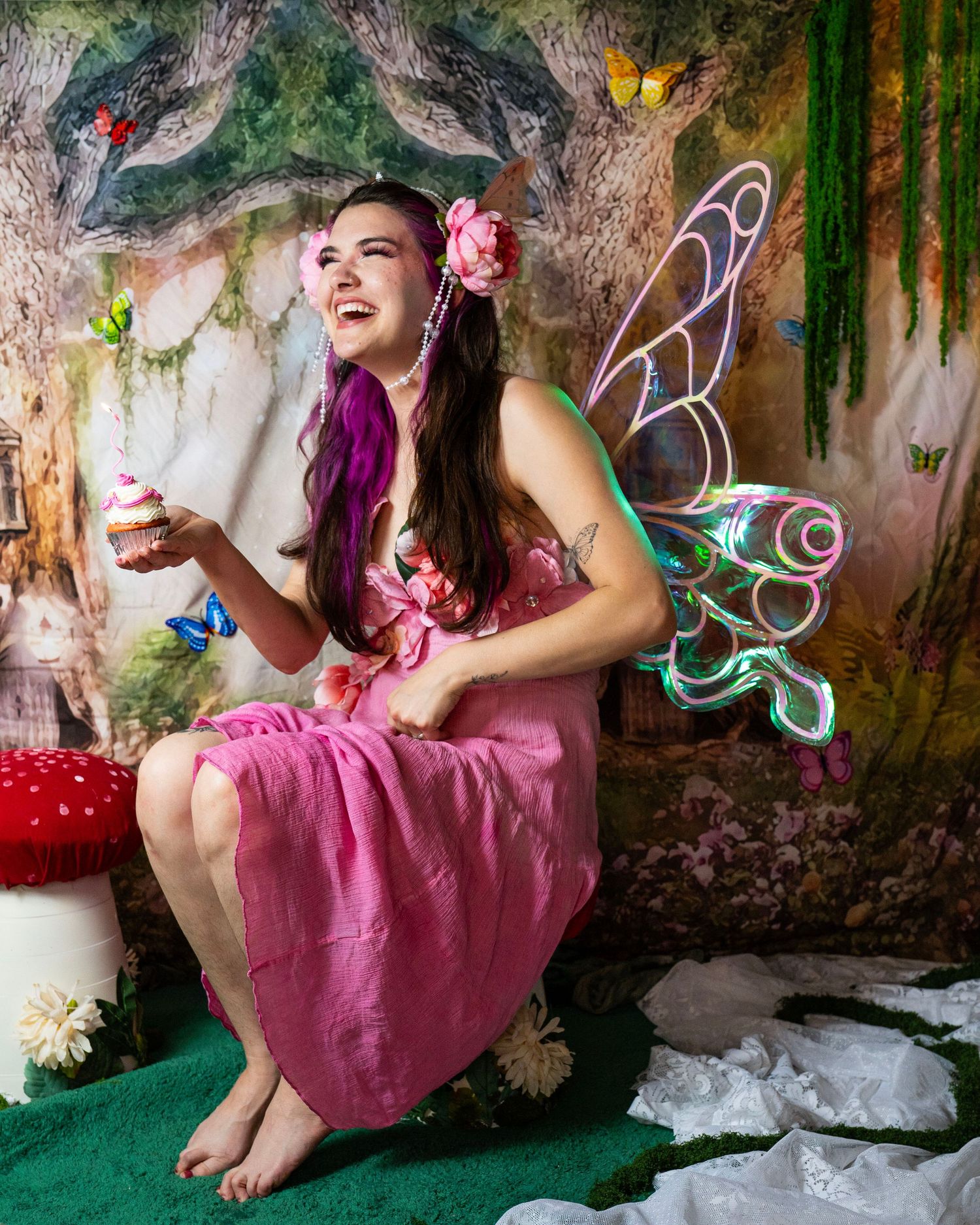Laughing fairy cosplay model in pink dress with LED wings sits in decorated enchanted forest setting. Fantasy Photoshoot