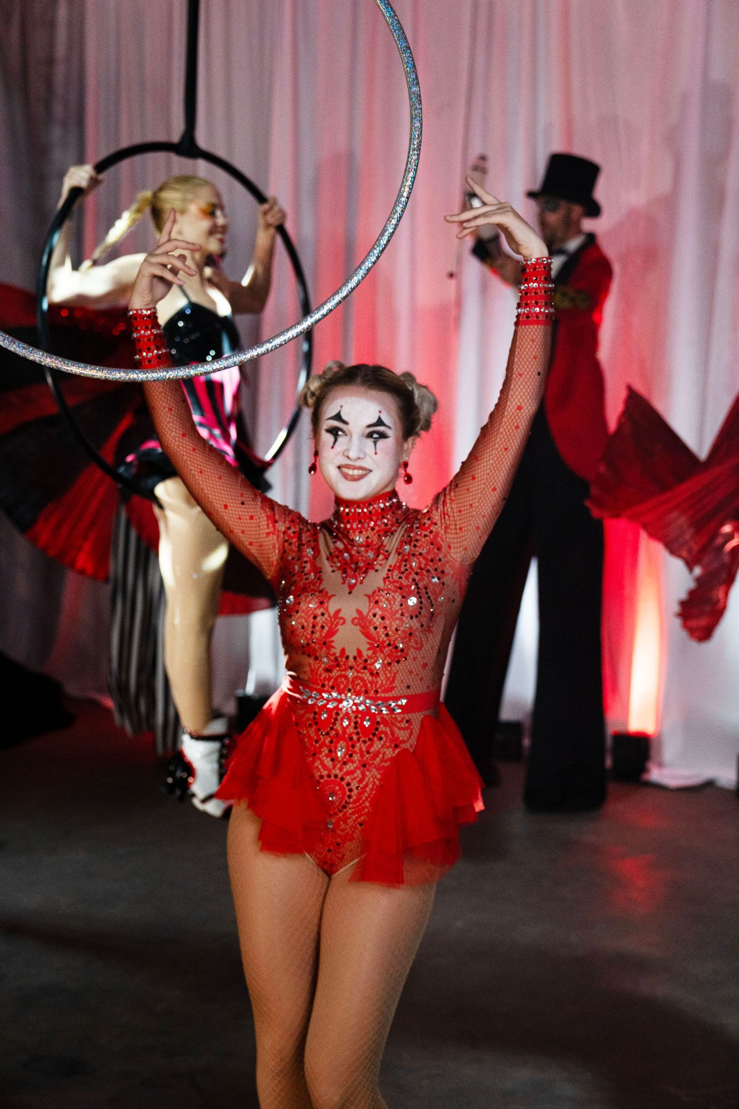 A smiling circus performer in a red sequined costume twirls a hula hoop at a circus-themed event.